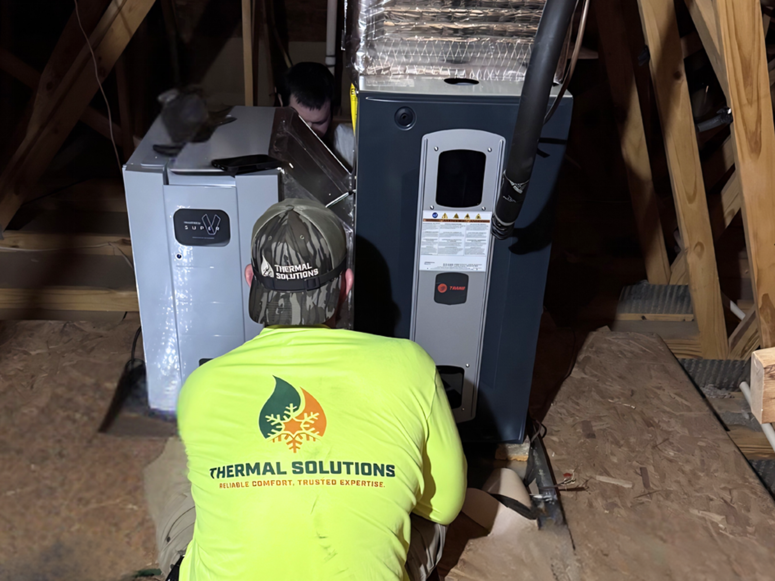 Two workers in yellow shirts with 'THERMAL SOLUTIONS' logo on back are installing or repairing HVAC equipment in an attic space with exposed wooden beams. One worker is kneeling while the other is standing.