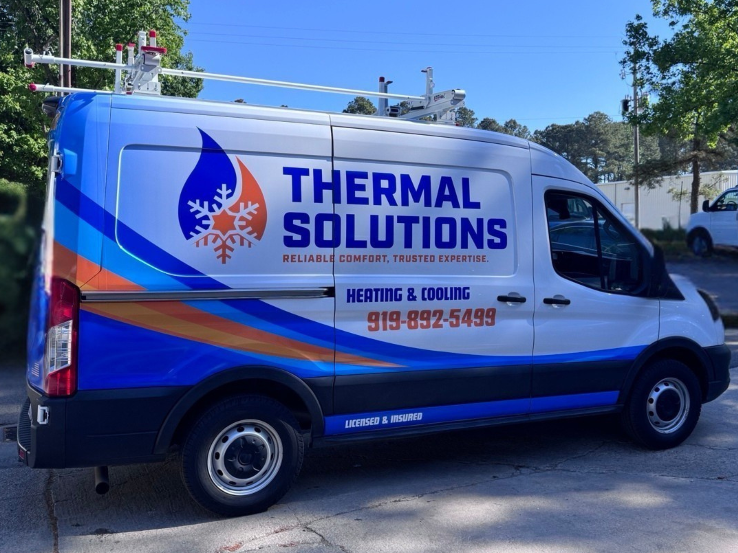 A white service van with a logo and contact information for Thermal Solutions, a heating and cooling company, parked outdoors with trees and a blue sky in the background.