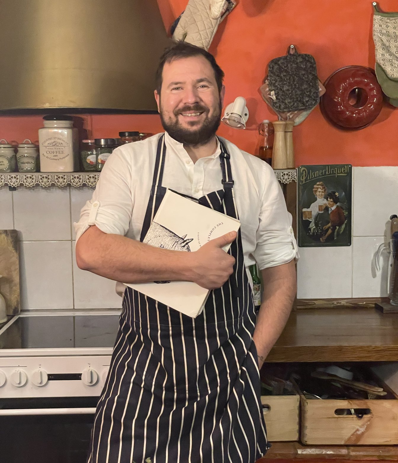A smiling man in a white shirt and striped apron holding a menu in a kitchen with orange walls, shelves with jars and kitchen utensils, and vintage-style decor.
