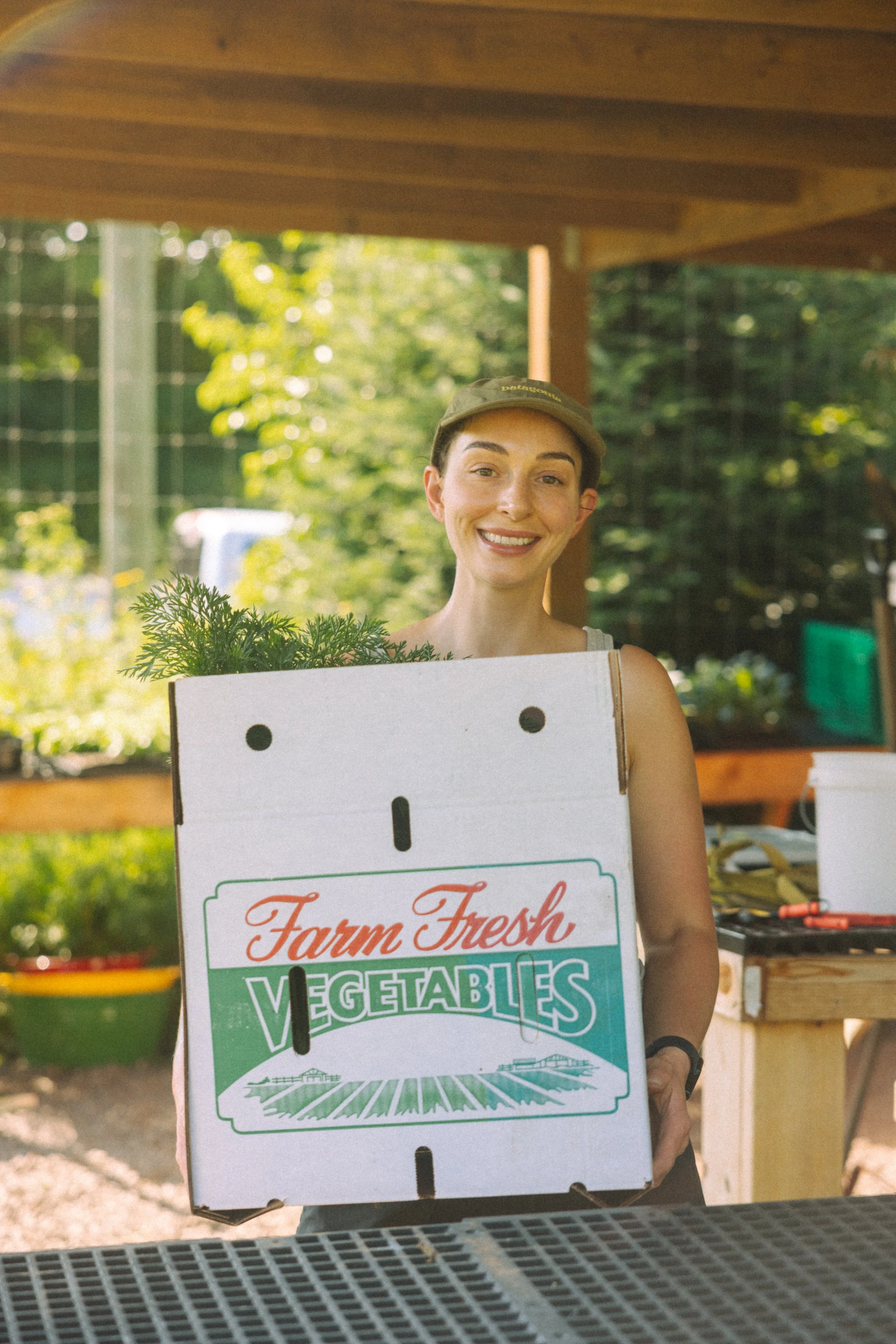 A woman smiling, holding a box of fresh vegetables and produce. She is wearing a khaki cap and tank top, with greenery in the background.