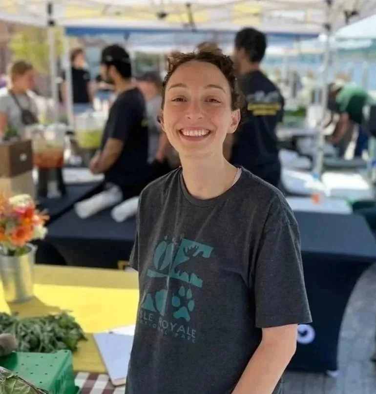 Smiling woman with short curly hair at outdoor market or food festival, wearing a dark T-shirt with a dinosaur graphic.