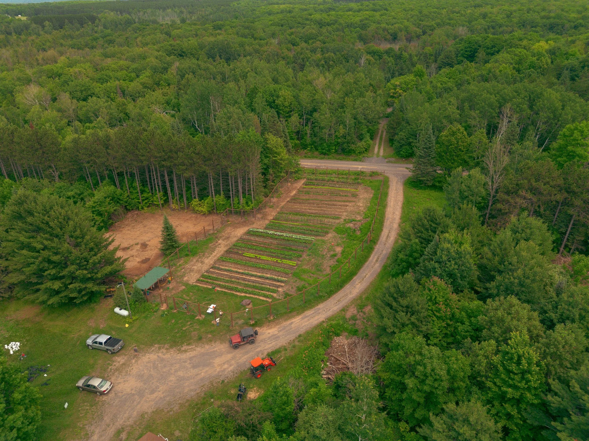 Aerial view of garden with planted rows of vegetables, surrounded by trees and woods, with a dirt driveway, parked trucks, and small structures.