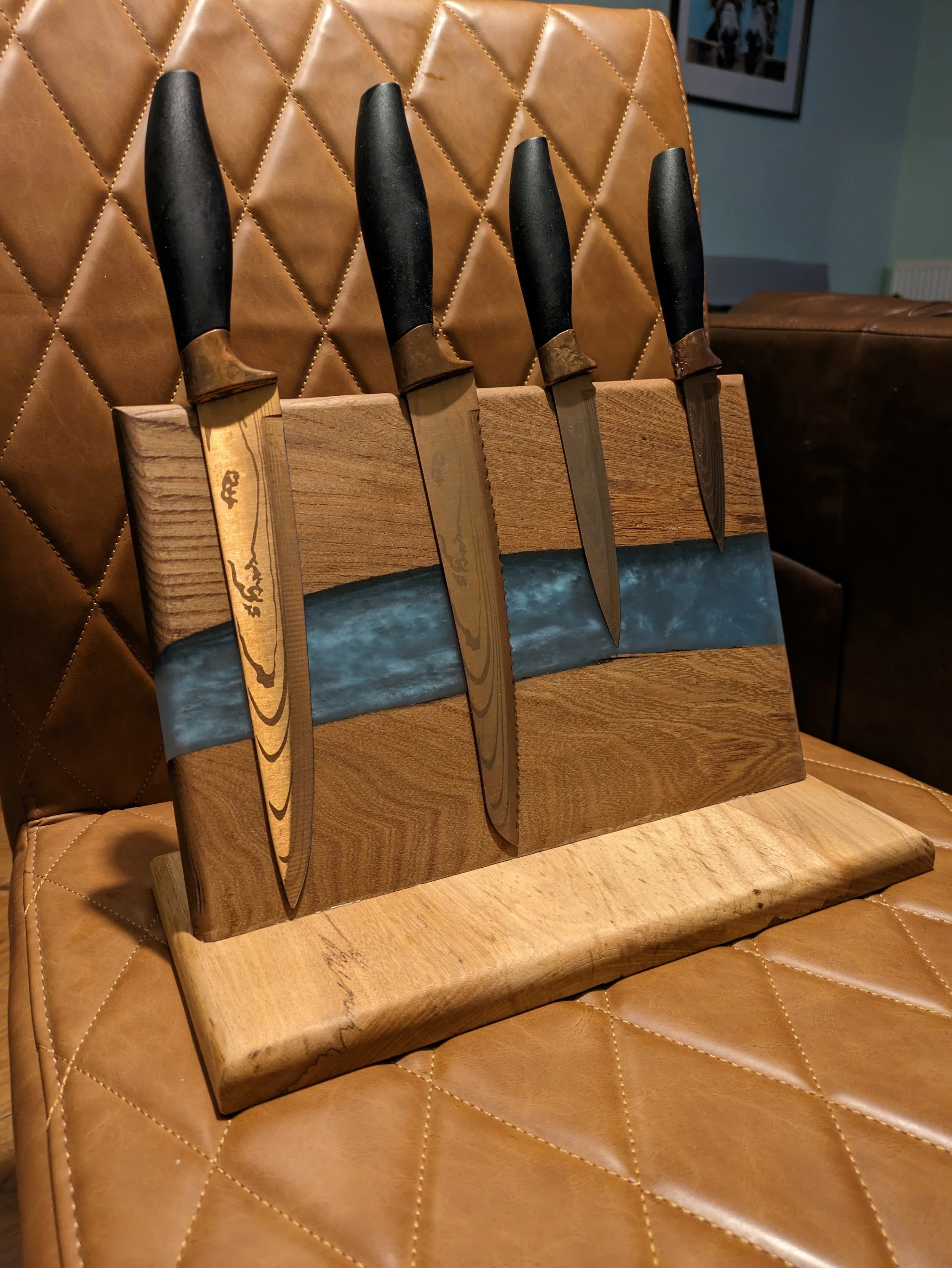 Four knives with black handles and metal blades inserted into a wooden knife holder on a leather-tufted chair.