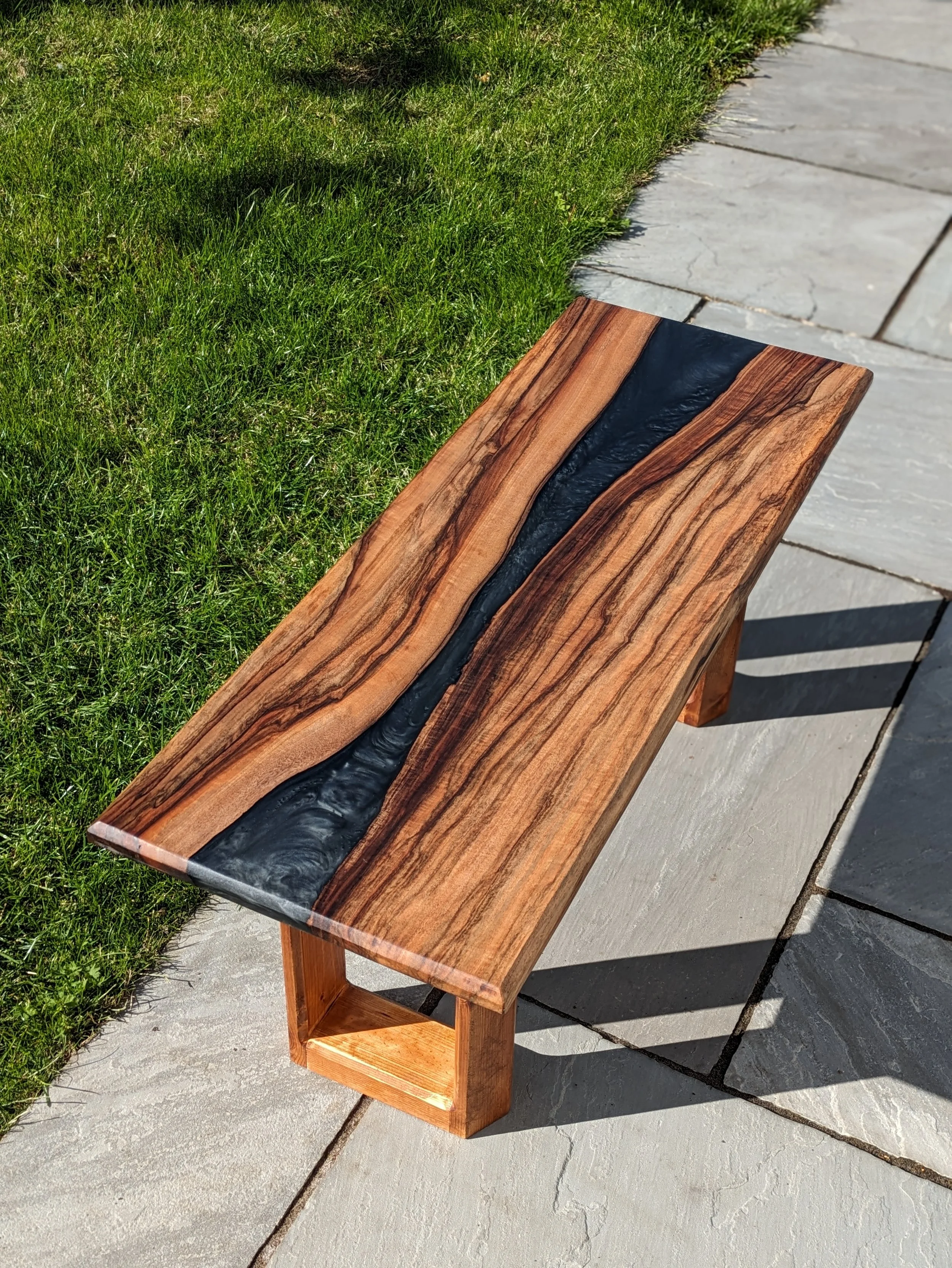 A wooden bench with a black resin river running through the center, placed on a concrete sidewalk next to a grassy lawn.