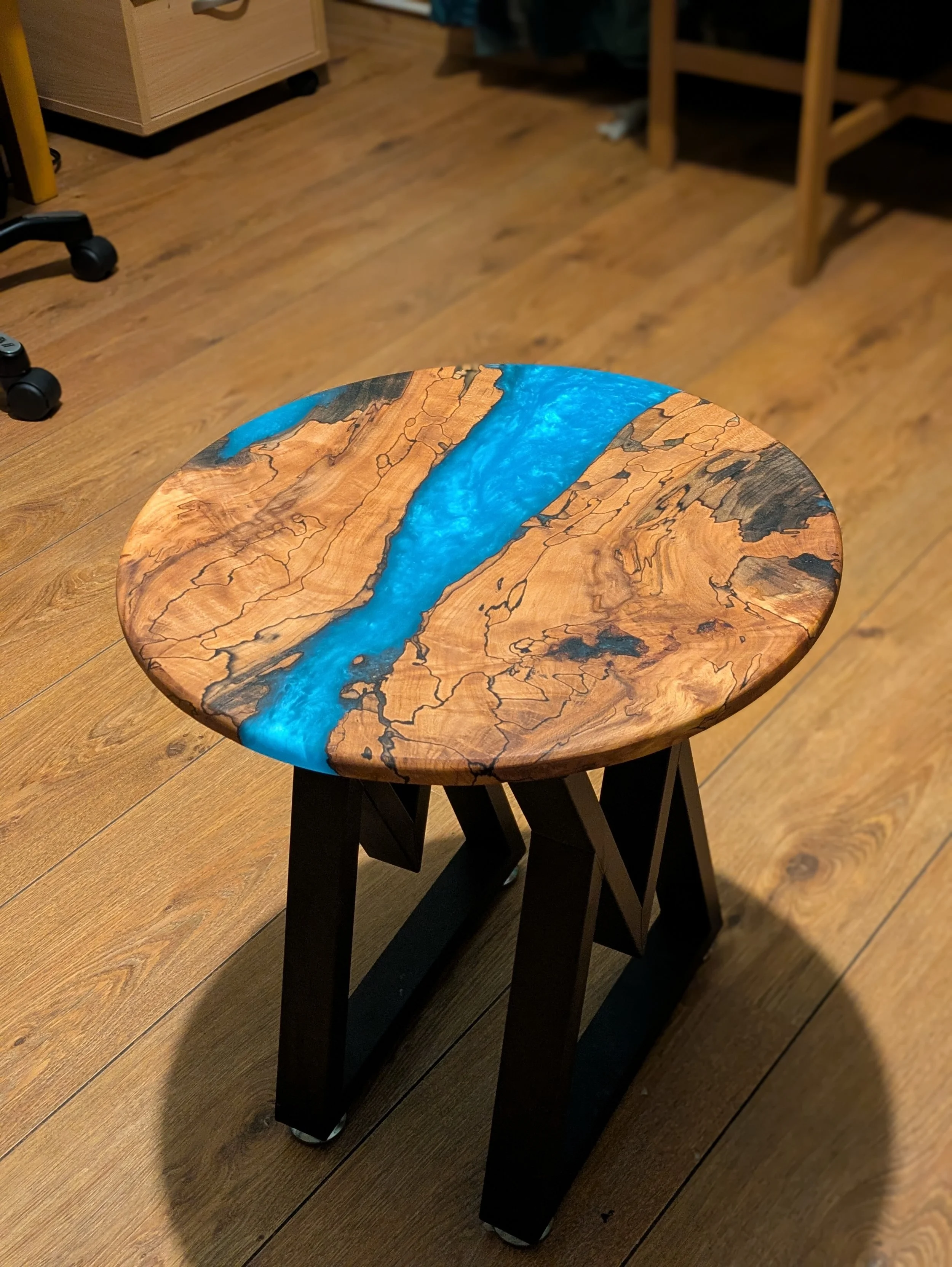 A wooden table with a blue resin inlay design on the top, situated on a wooden floor in a room with furniture.