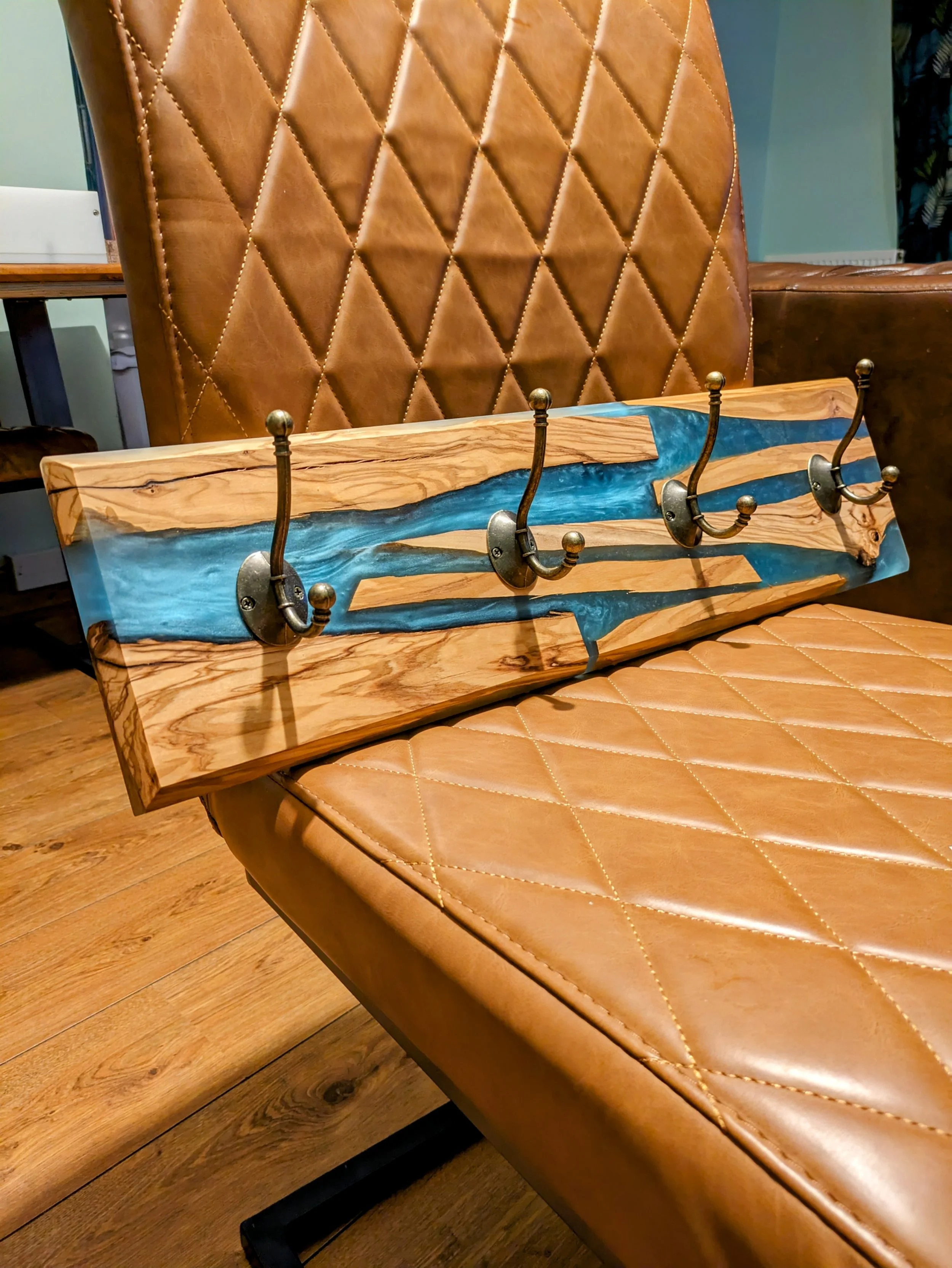 A wooden coat rack with four hooks in front of a brown leather chair with a diamond quilted pattern. The background includes a table and wall with a radiator.