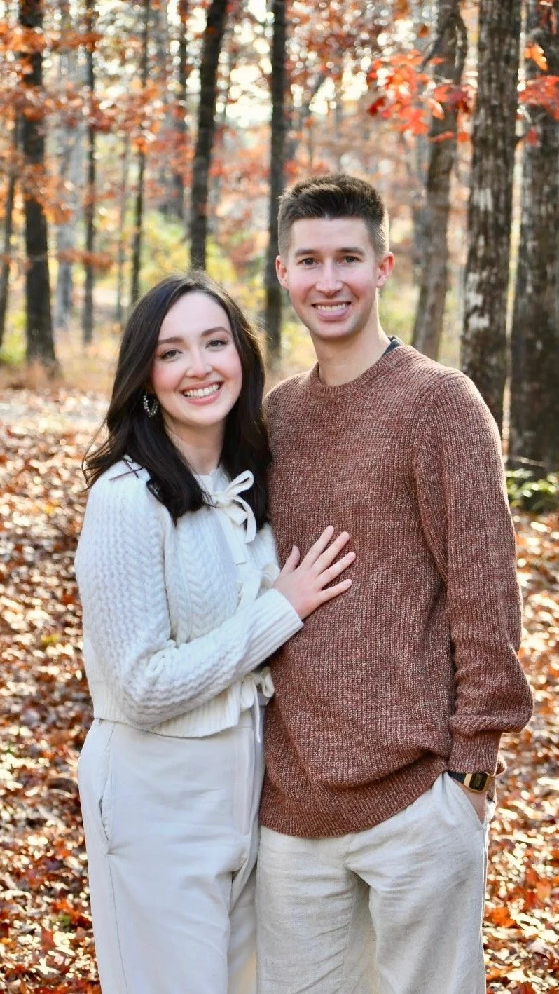 A smiling young couple standing together in a forest with autumn leaves, the woman wearing a white sweater and the man in a brown sweater, during daytime.