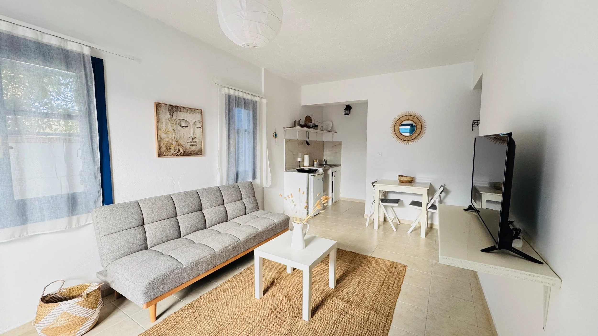 Living room with a grey sofa, white coffee table, TV on white console, framed artwork of a face, small dining table with two chairs, open kitchen area, and decorative mirror on white walls.