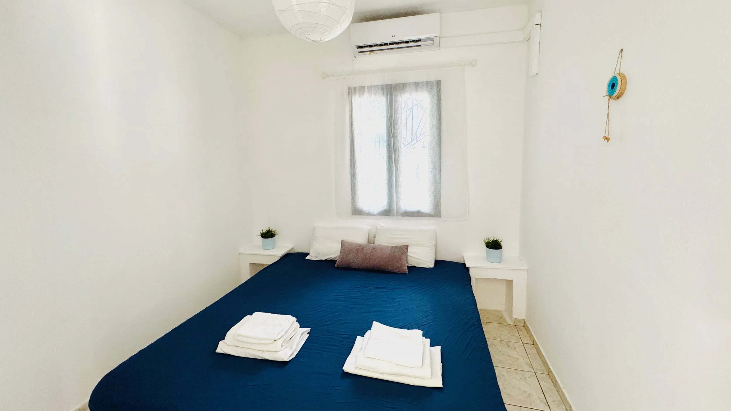 A minimalist bedroom with a blue bedspread, white pillows, and folded towels on the bed. There are two small nightstands with potted plants on each side of the bed, a window with sheer curtains above the headboard, and a wall-mounted air conditioner 