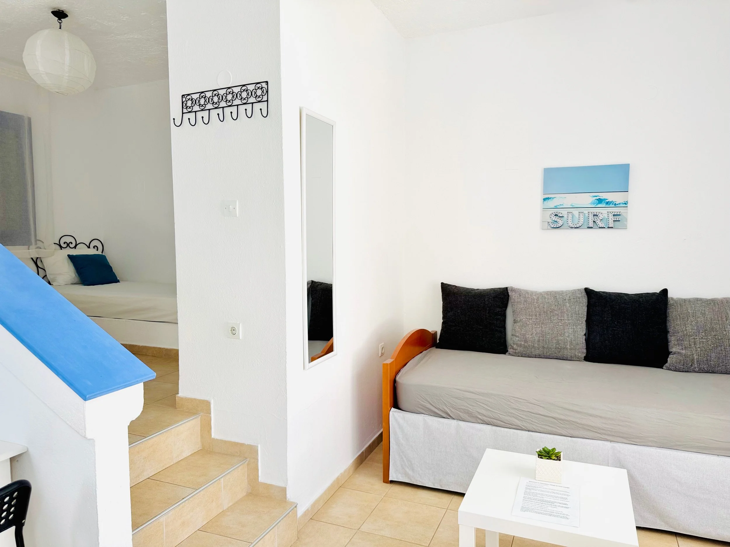 A living room with a beige sofa, black and gray pillows, a white coffee table with a small potted plant, a wall painting with the word 'SURF', and a mirror on the wall. There is an open doorway leading to a bedroom with a bed and blue pillows, and a 