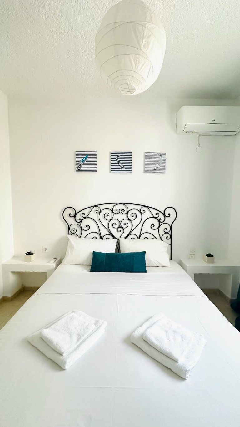 A neatly made bed with a decorative wrought iron headboard, white sheets, and teal pillows. Two folded white towels are placed at the foot of the bed. The room features a white hanging paper lantern, three framed pictures above the bed, and small pla