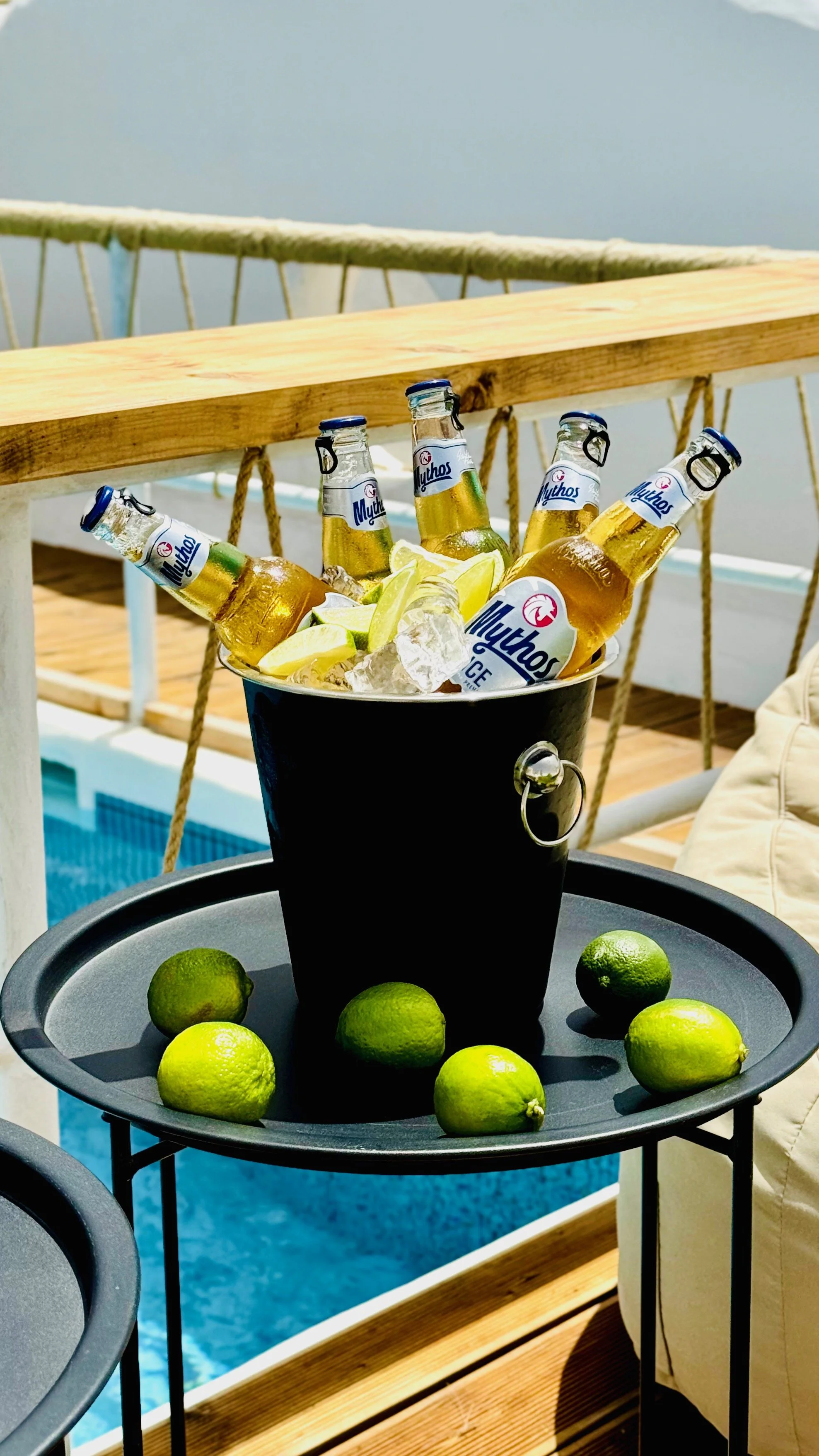 Bucket of beer bottles and lemon wedges with limes on a tray, set on a balcony overlooking a pool.