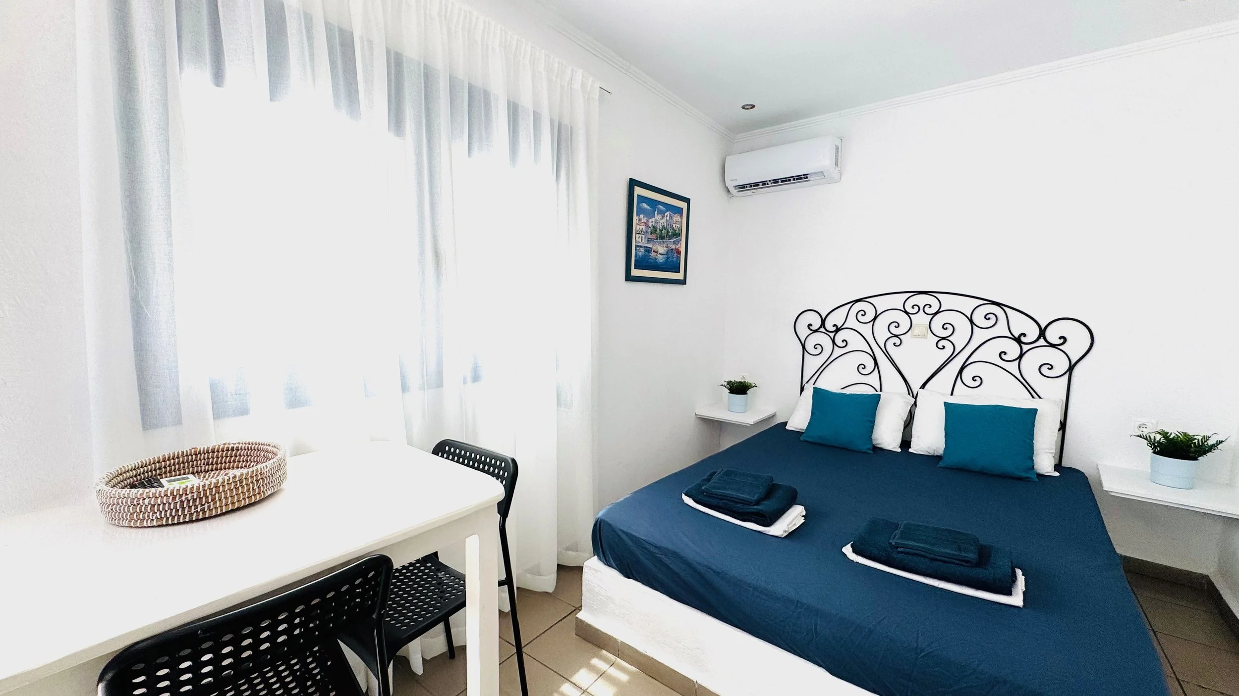 Apartment with a blue bed, white walls, black wrought iron headboard, white side tables with potted plants, a painting on the wall, sheer curtains, and a white desk with black chairs.