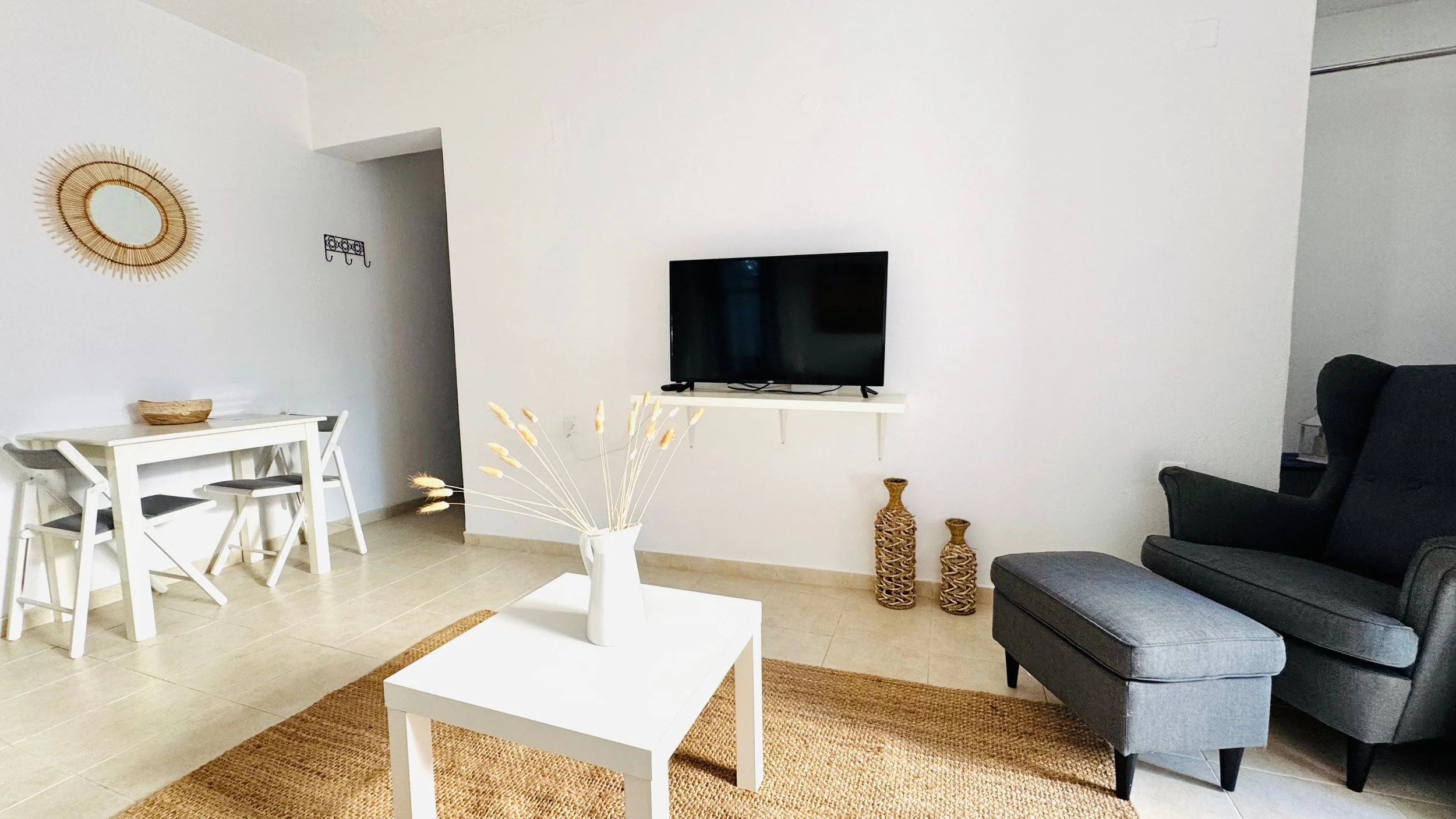 Living room with white walls, black armchair and ottoman, wall-mounted TV, white coffee table with vase and dried plants, beige rug, white dining table with chairs, decorative woven vases, and wall decor.