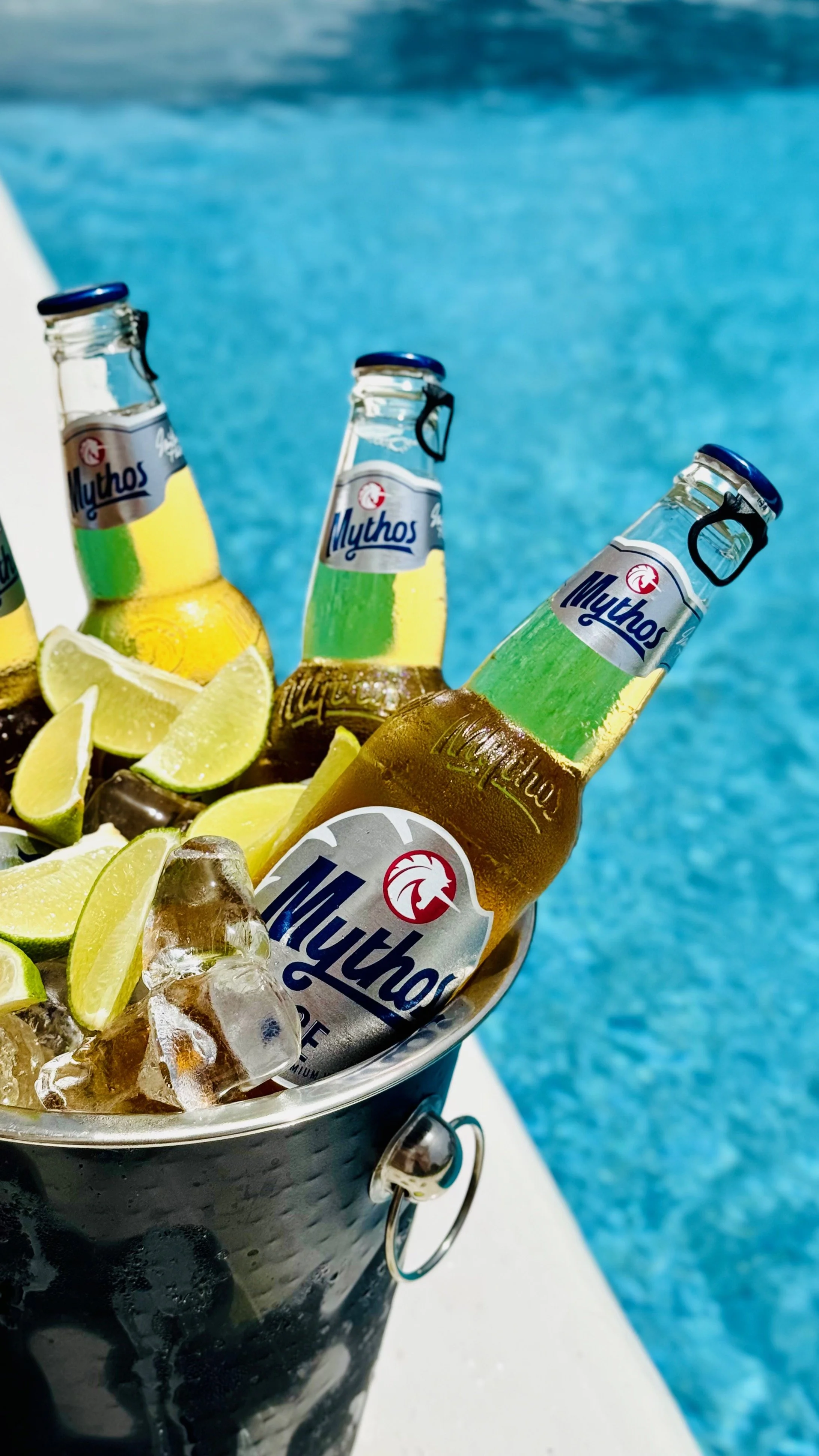 A black metal bucket filled with ice, lemon wedges, and three bottles of Mythos beer near a swimming pool.