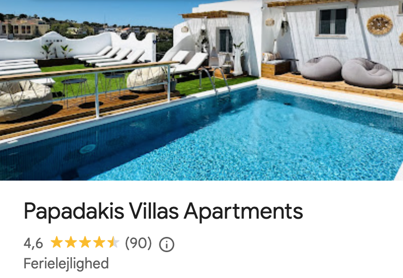 Outdoor pool area at Papadakis Villas Apartments with lounge chairs, bean bag chairs, and a white building with a deck and small yard.