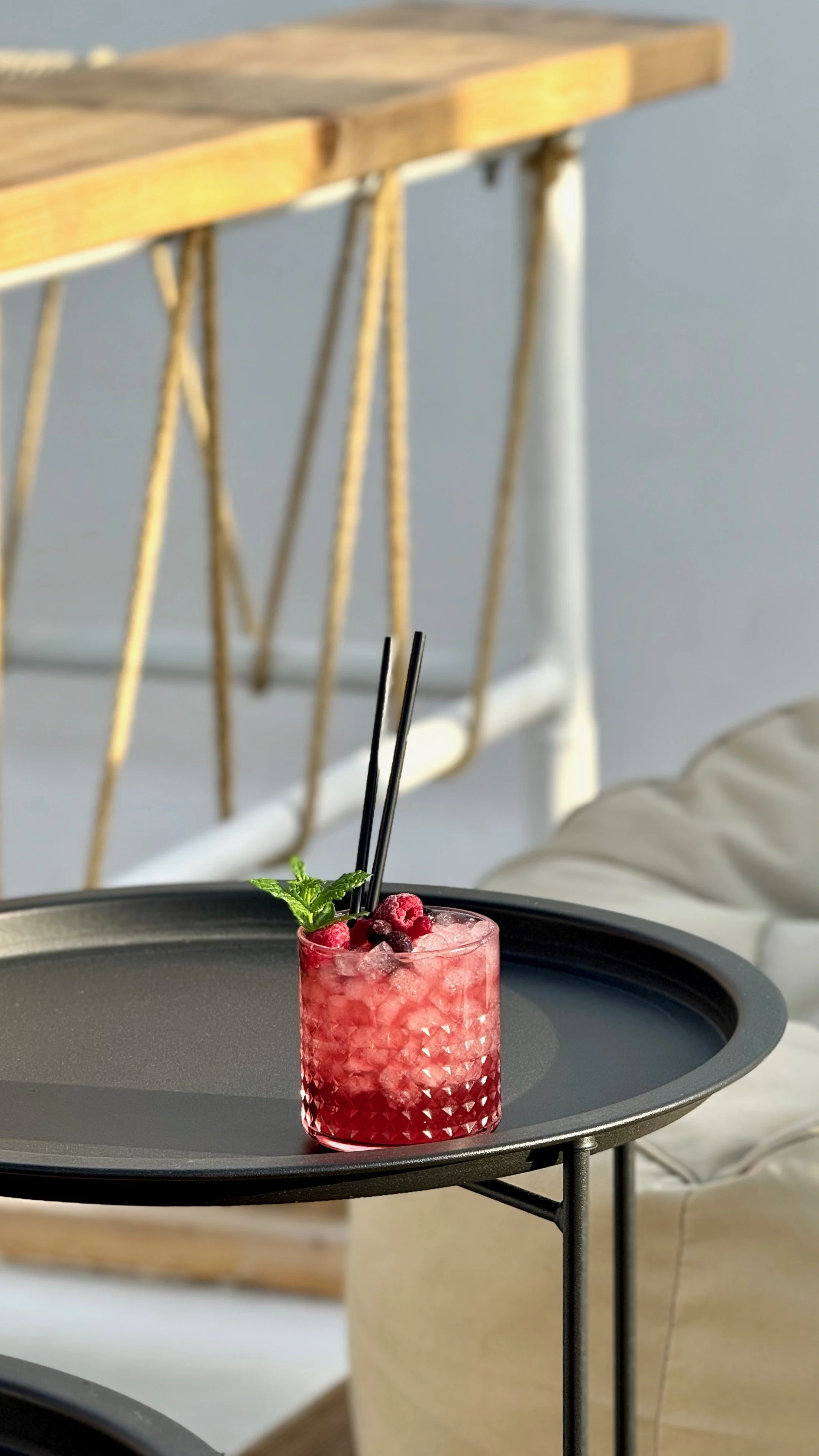 A pink cocktail with ice, garnished with berries and a mint sprig, on a black tray.