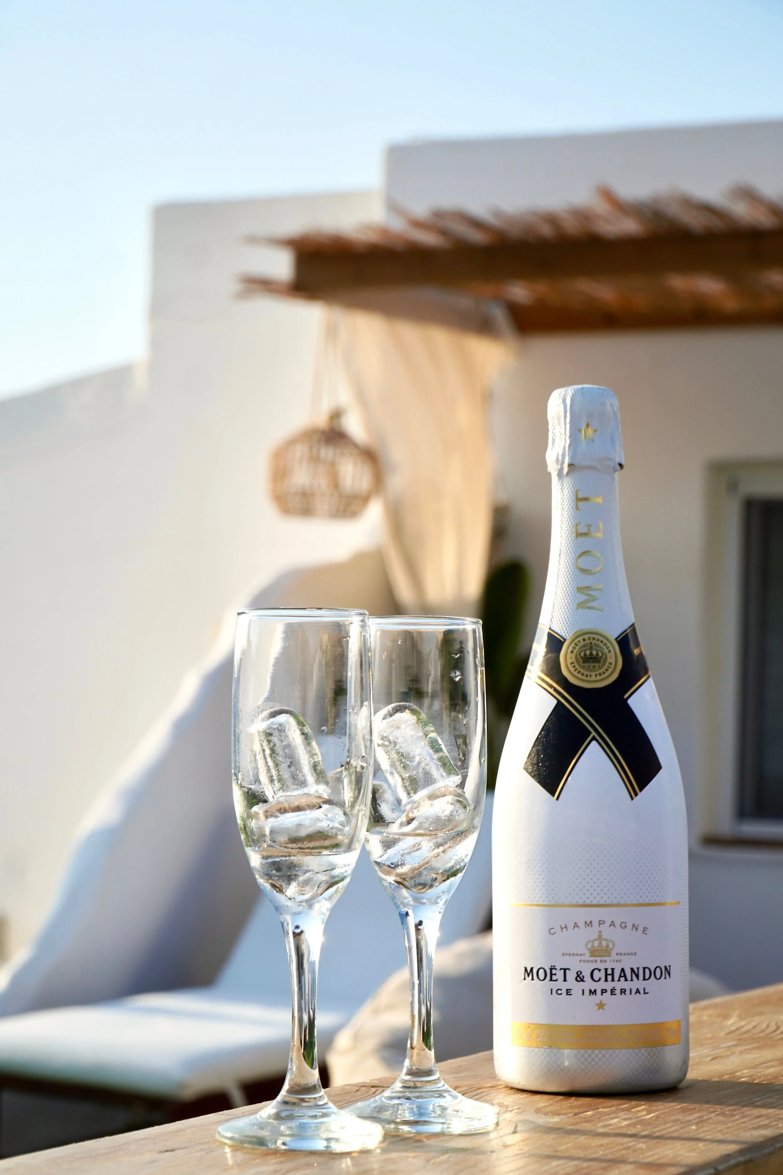 A bottle of Moët & Chandon Champagne and two glasses of champagne with ice on a wooden table outdoors, with a sunny sky and white building in the background.