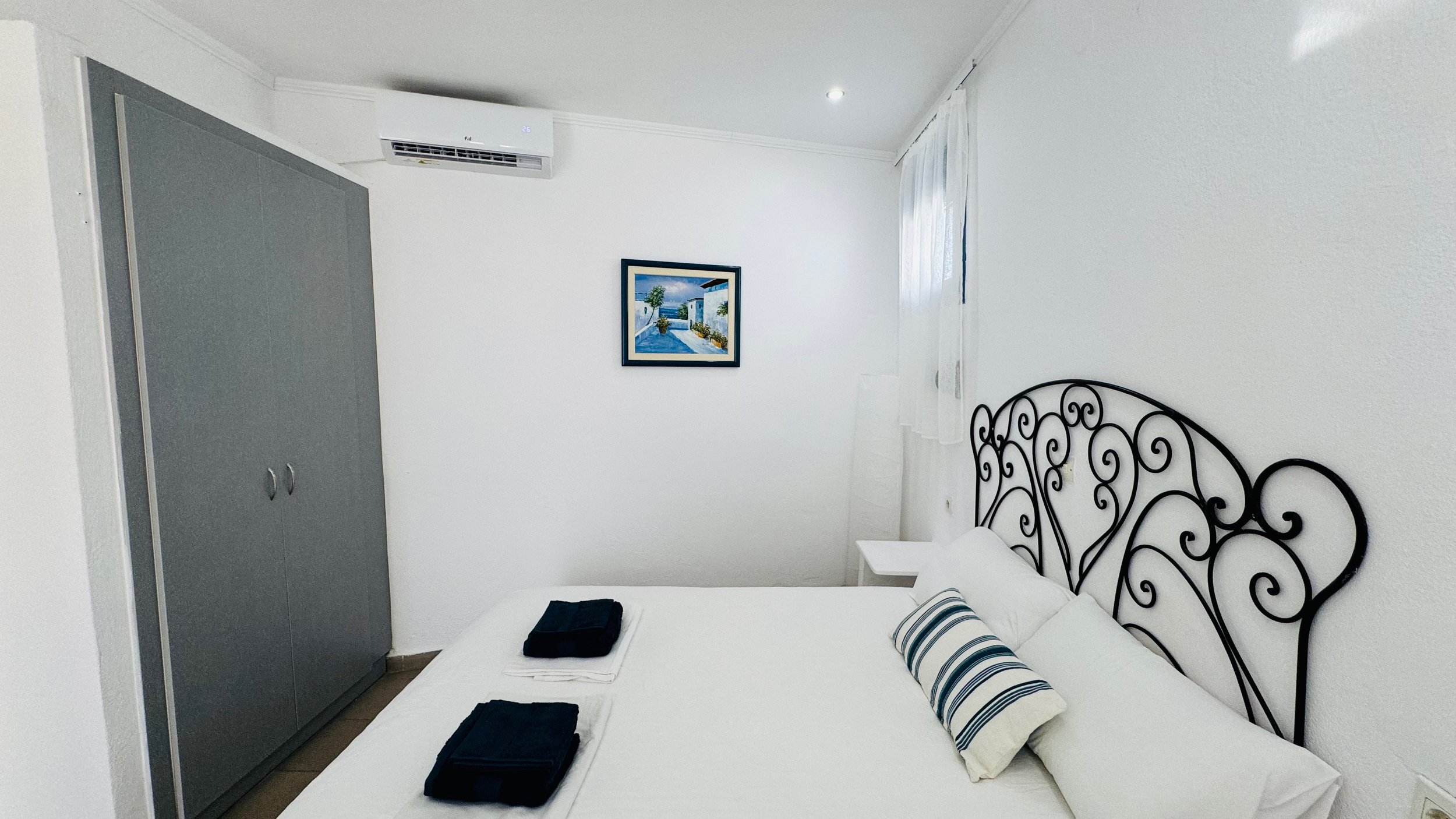 Minimalist bedroom with a double bed, ornate black metal headboard, white bedding, striped pillow, folded towels, large gray wardrobe, wall-mounted air conditioner, and framed picture on the wall.