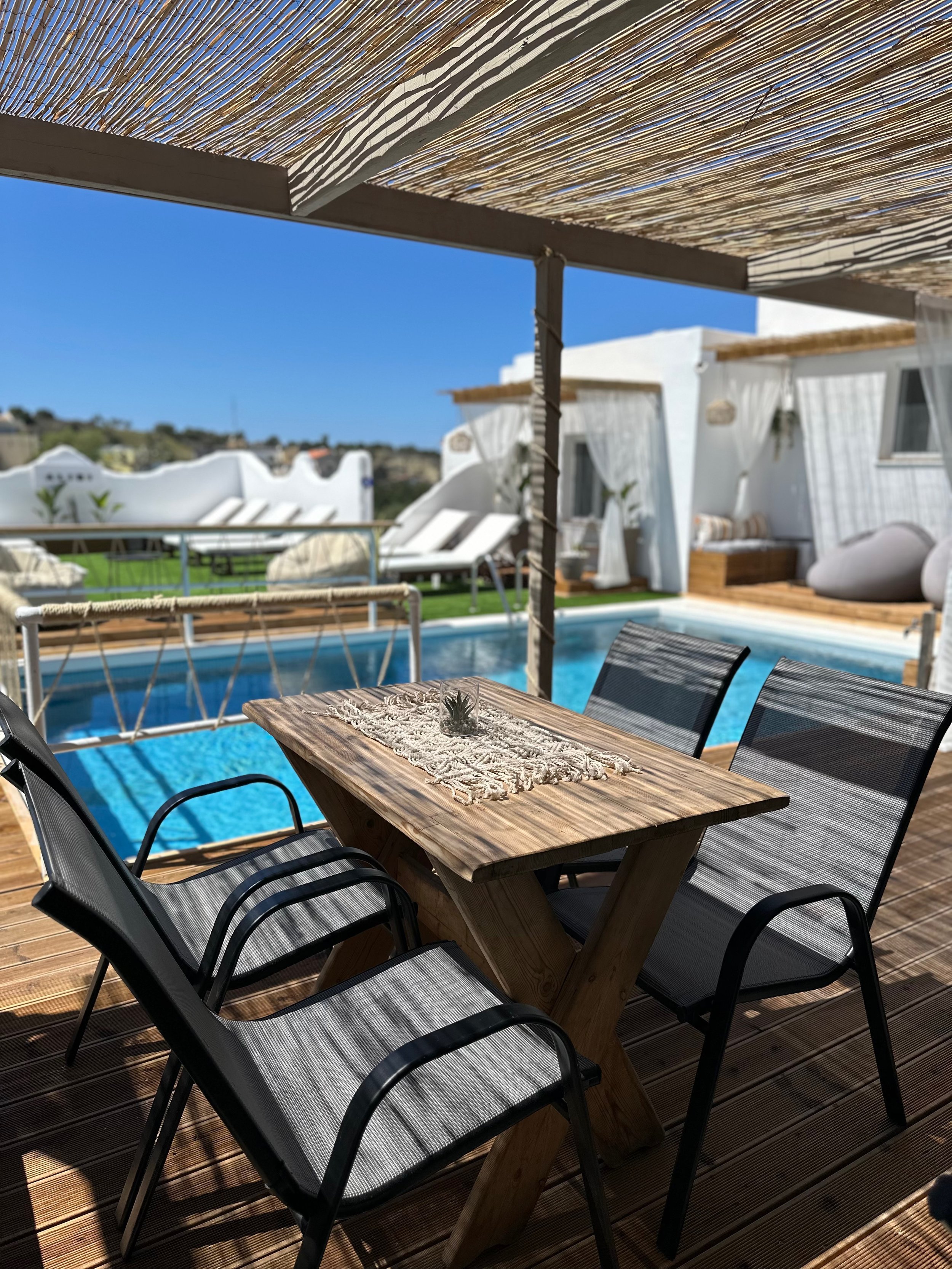 Outdoor patio with wooden table and chairs next to a pool, covered by a bamboo pergola, with white buildings and deck chairs in the background.