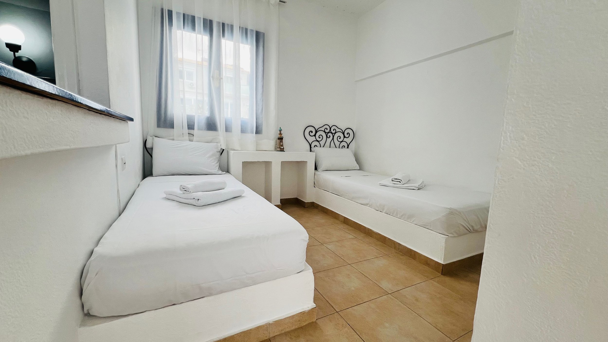 Minimalist bedroom with two single beds, white sheets, and a window with sheer curtains, featuring a decorative headboard and tiled floor.