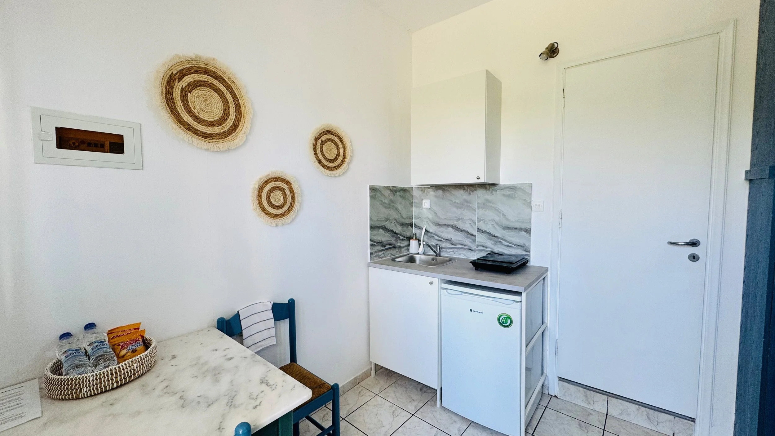 Small kitchen with a white mini fridge, a sink, a two-burner electric stove, and white cabinets. Decor includes three brown, concentric circle wall hangings. A table has water bottles and snacks, and a blue chair is at the table.