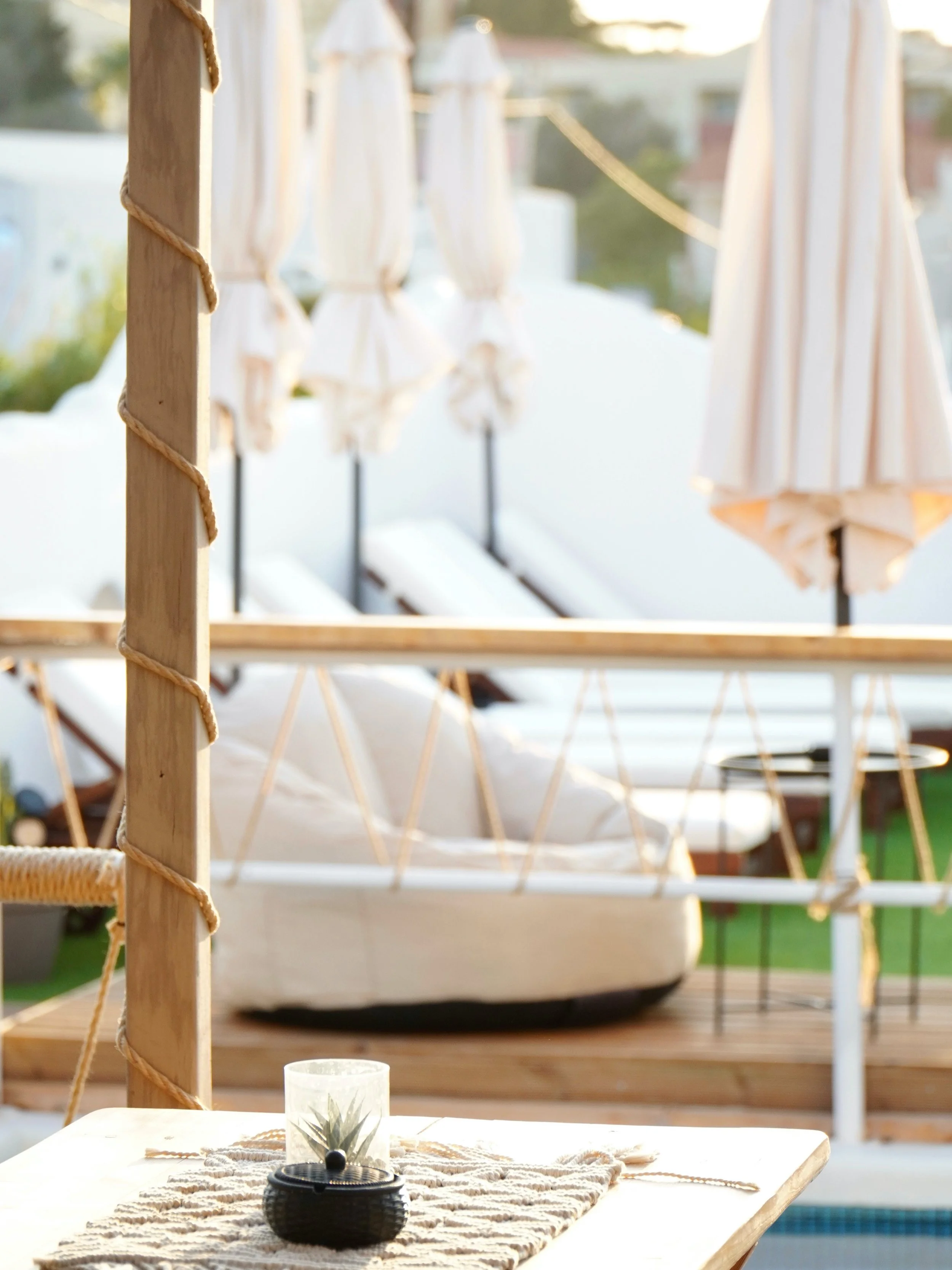 A cozy outdoor patio area with a table, candle, and plant decoration. In the background, there are sun loungers with umbrellas and a boat docked nearby, all illuminated by warm outdoor lighting.