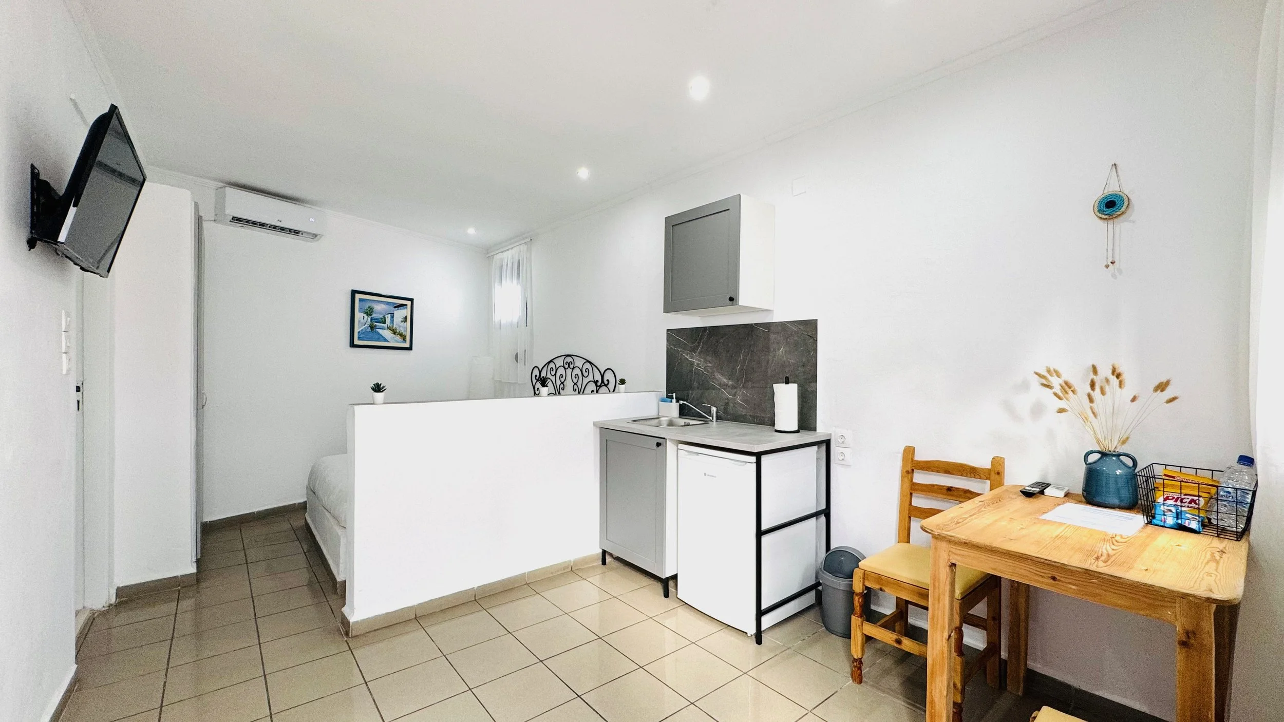 Small studio apartment with white walls and beige tiled floor, featuring a wall-mounted TV, compact kitchenette with a sink, white and gray cabinets, black countertop, and a brown wooden dining table with a vase of dried flowers, a remote, a water bo