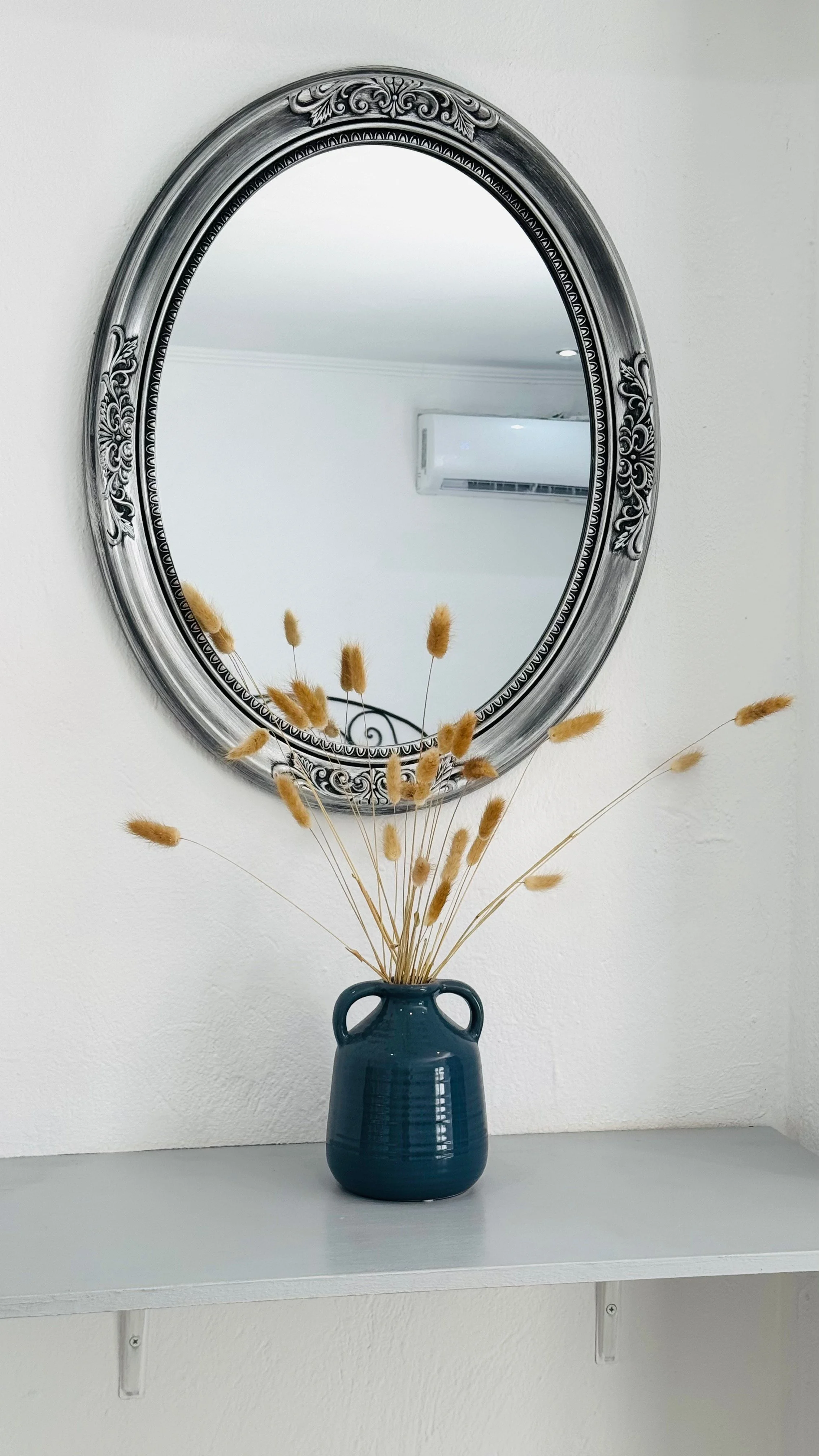 A decorative blue vase with dried grasses is placed on a white shelf beneath an ornate silver oval mirror on a white wall, with a reflection of an air conditioning unit.