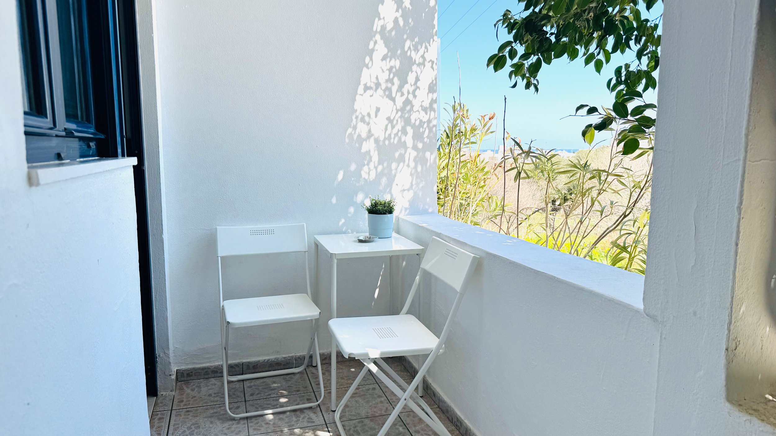 Small balcony with two white folding chairs, a white table, and a potted plant; sunny outdoor view with leaves and trees.