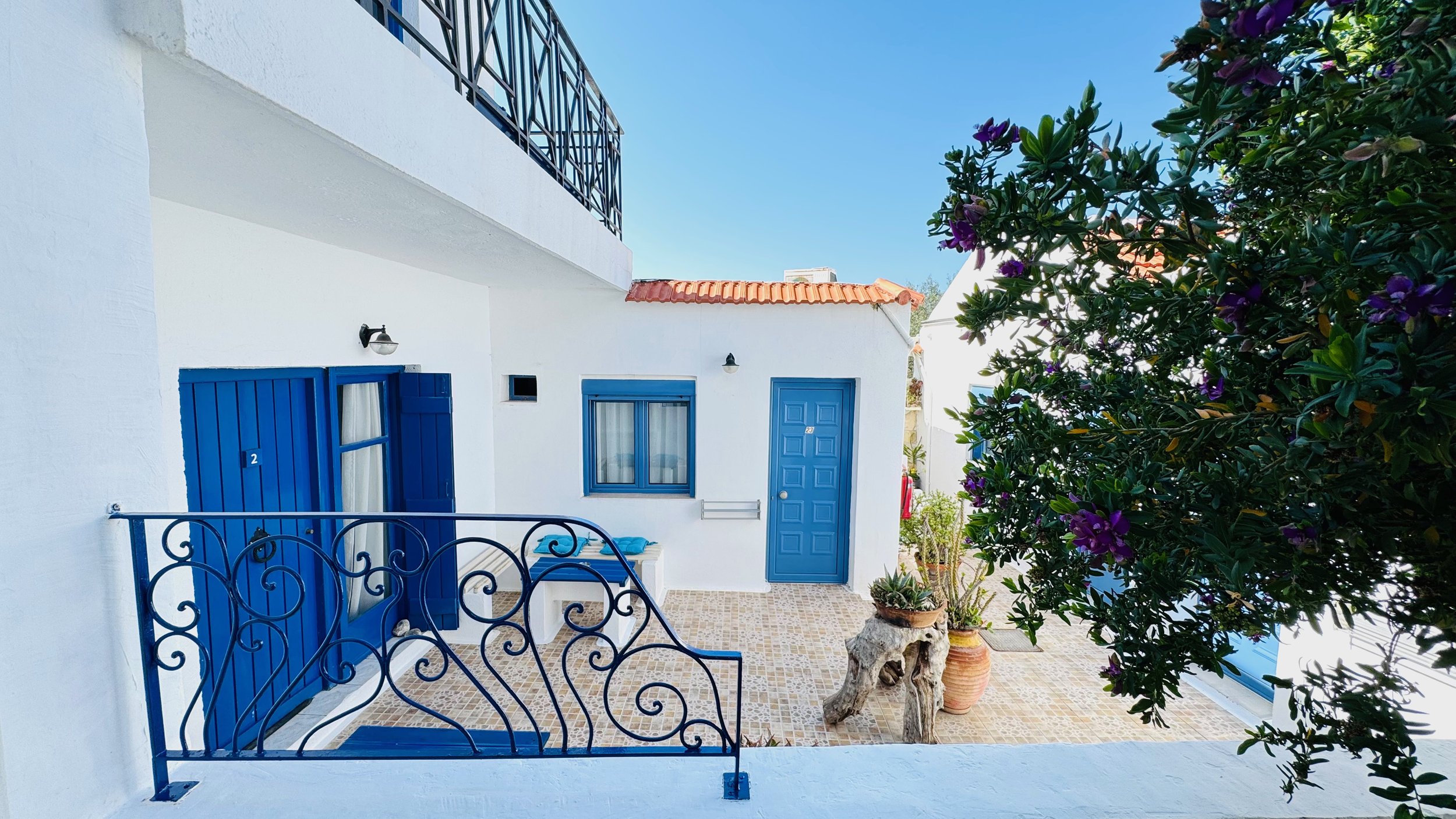 White Mediterranean-style area with blue doors and shutters, a tiled patio, potted plants, and purple flowering shrubs under a clear blue sky.