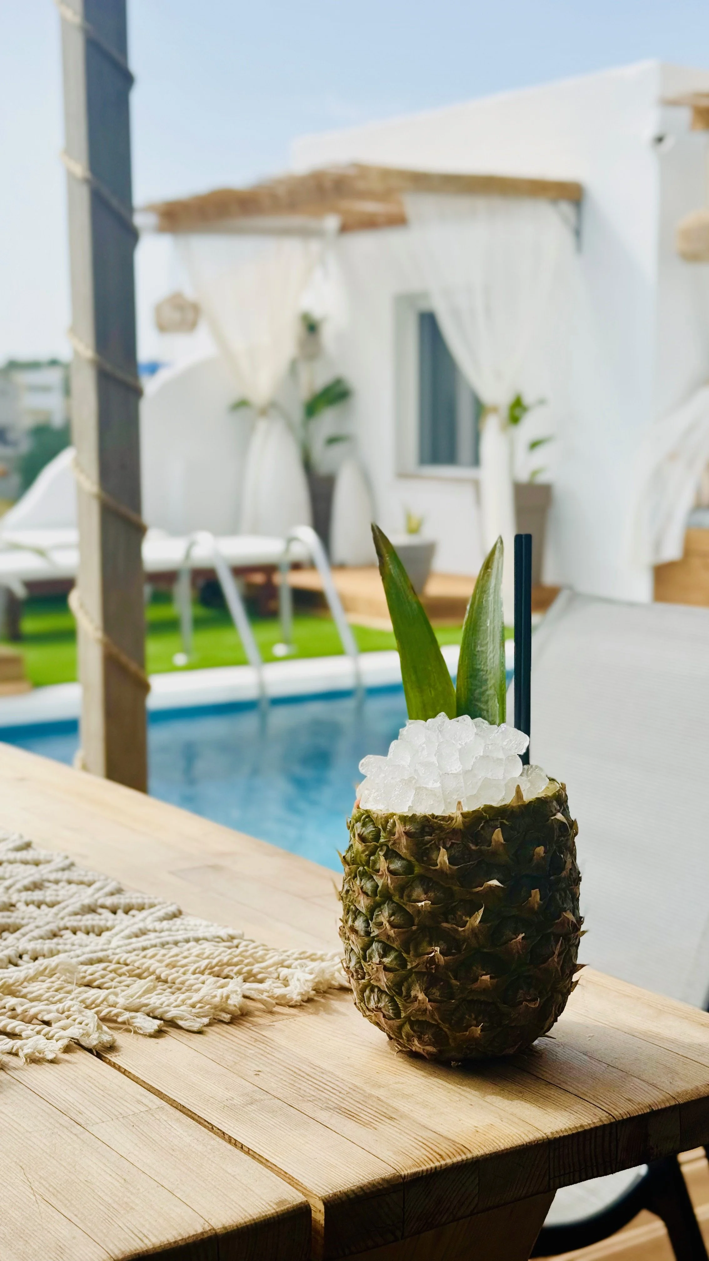 A drink served in a hollowed-out pineapple with two green pineapple leaves, crushed ice, and a straw on a wooden table with a macramé placemat. In the background, there is a pool, white curtains, and white buildings, suggesting a tropical outdoor set