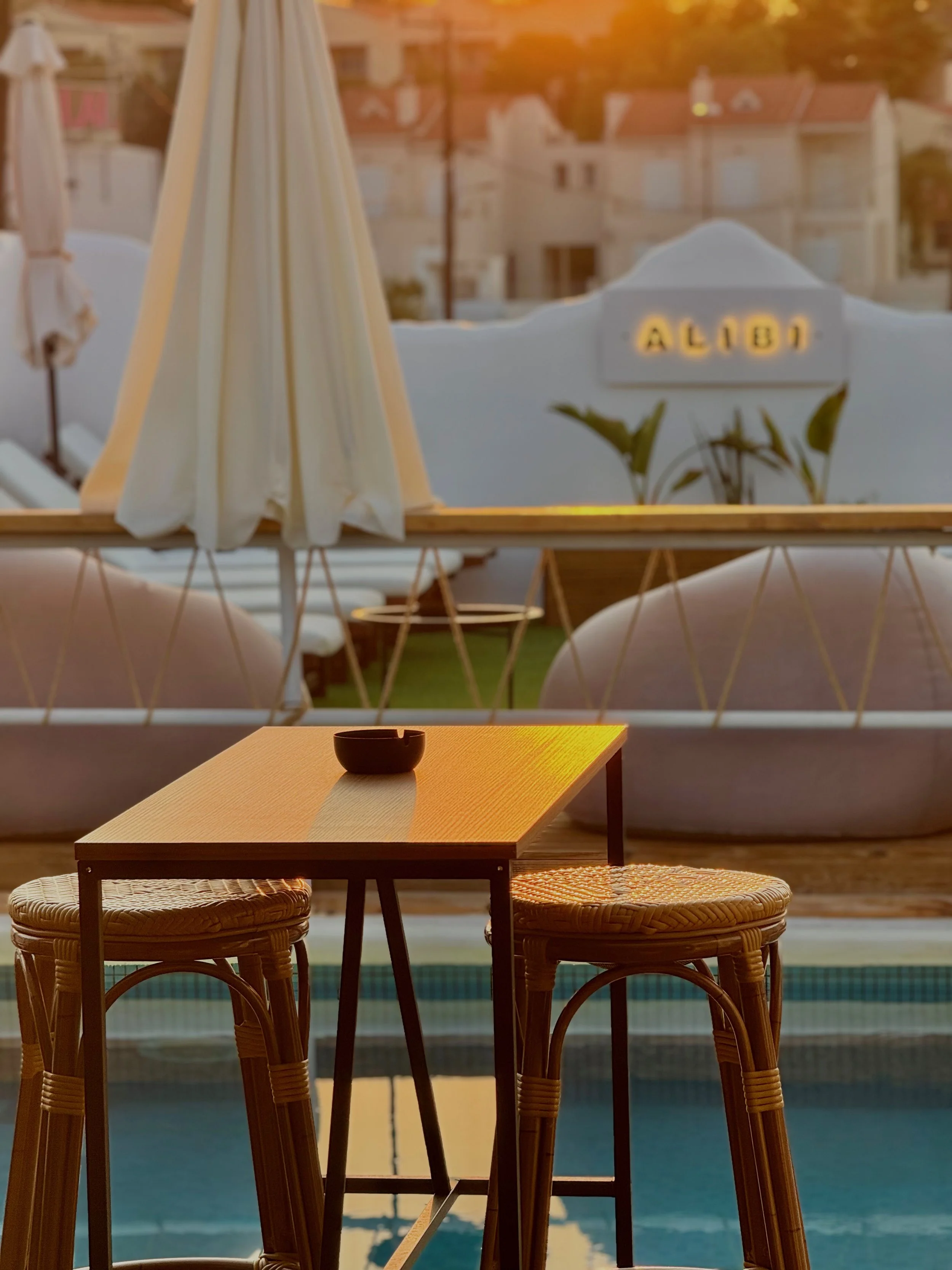 A cozy outdoor seating area at sunset featuring a small wooden table with two wicker chairs, a black ashtray on the table, and an outdoor pool in the background. There are white umbrellas and lounge chairs behind the table, with a blurred building an