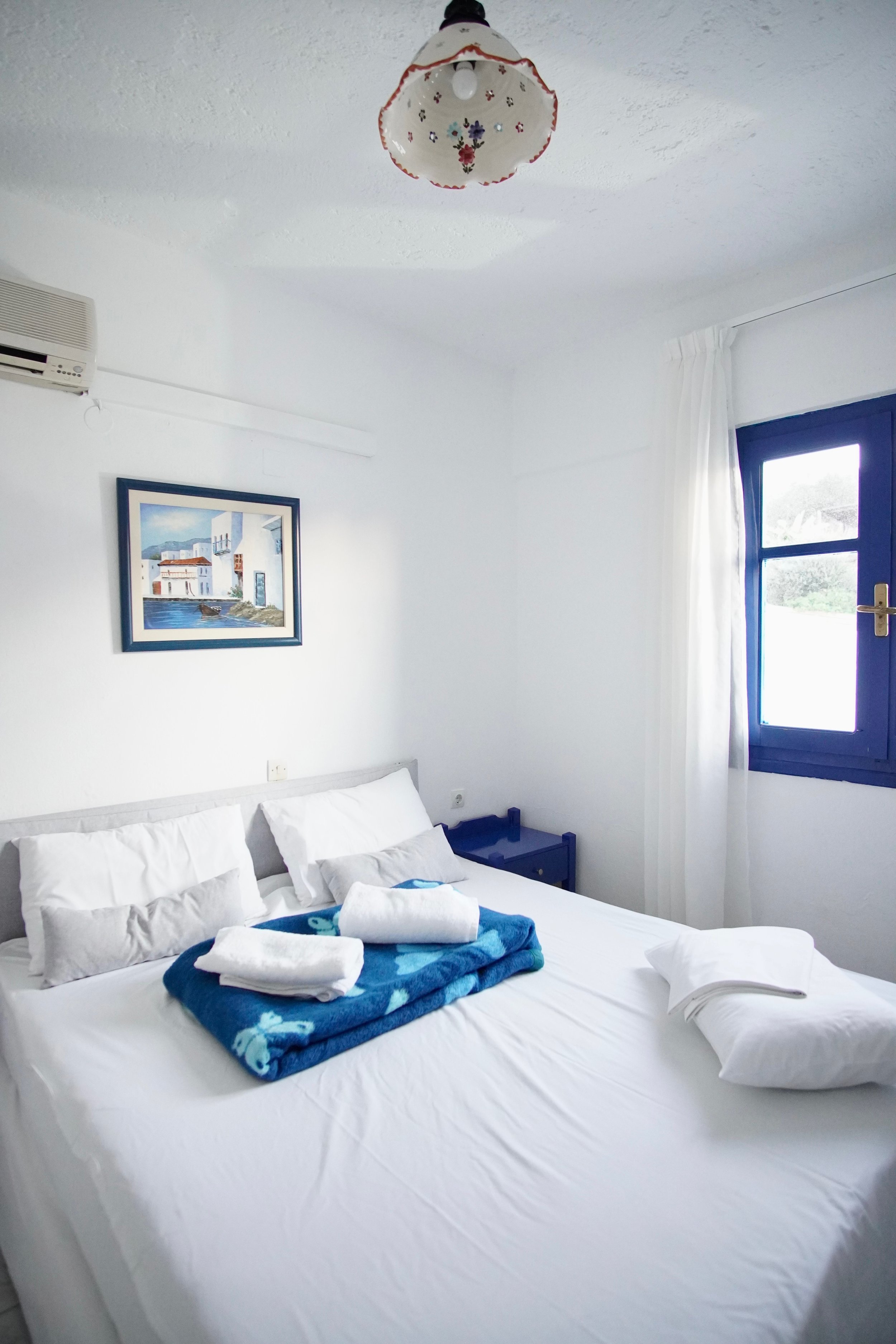 Cozy bedroom with a white bed, blue window, framed wall art, and overhead light.