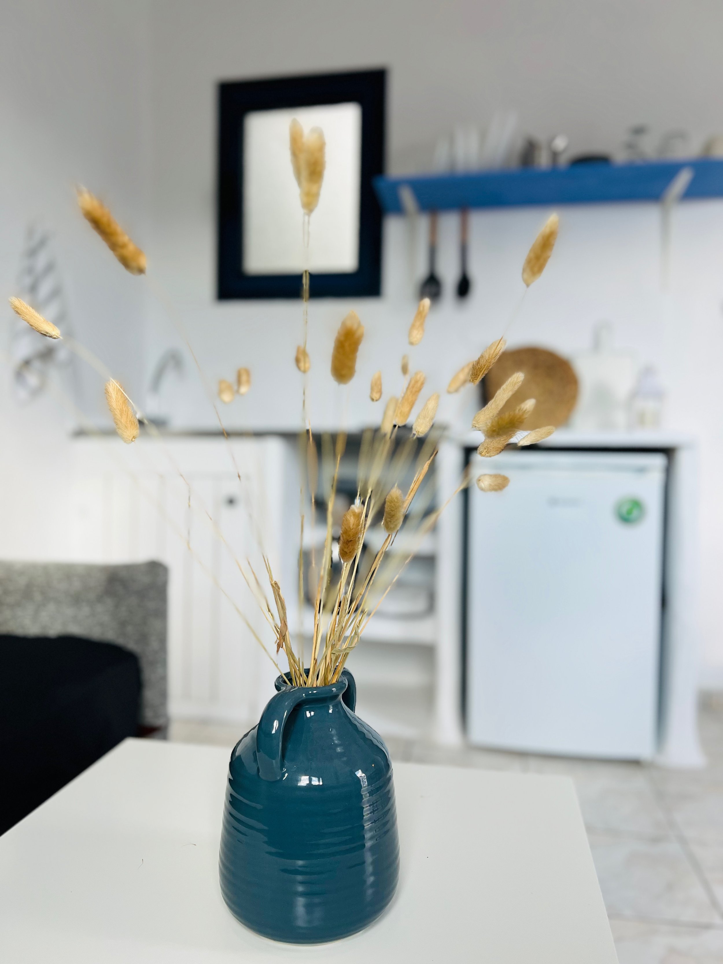 Decorative dried grasses in a blue vase on a white table, with a kitchen background featuring shelves, utensils, and a small refrigerator.