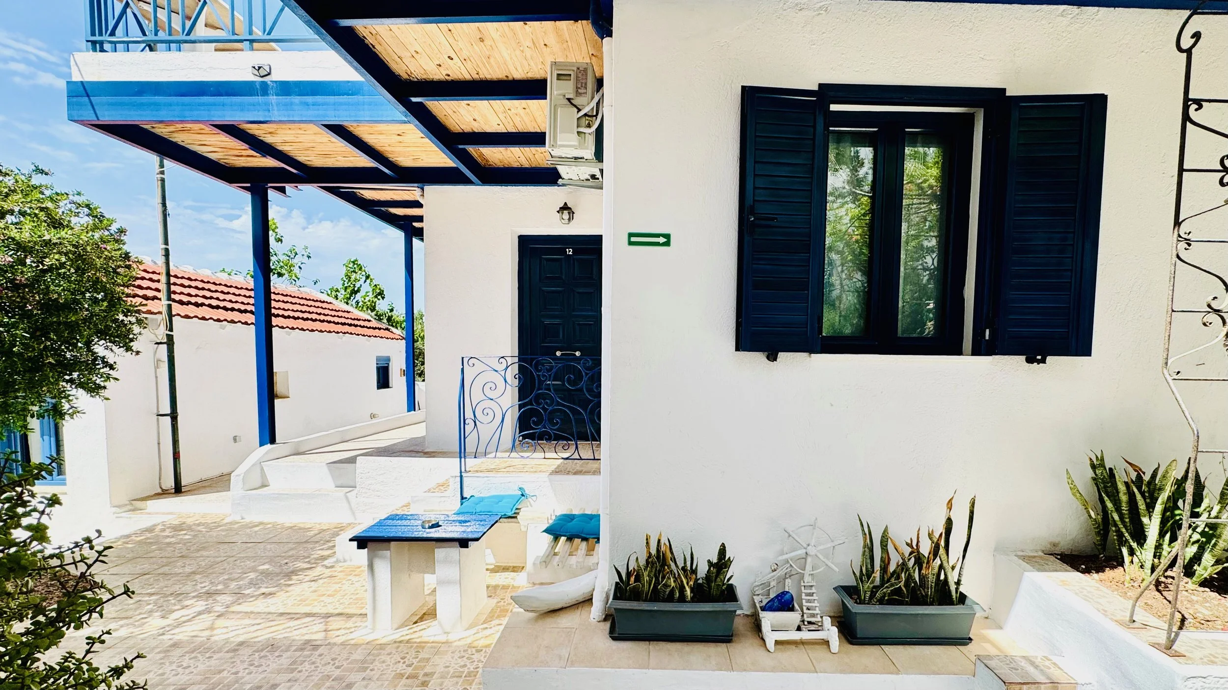 Exterior of a white Mediterranean-style house with a black door, black window shutters, and potted plants. There is a small porch with outdoor furniture and blue accents.
