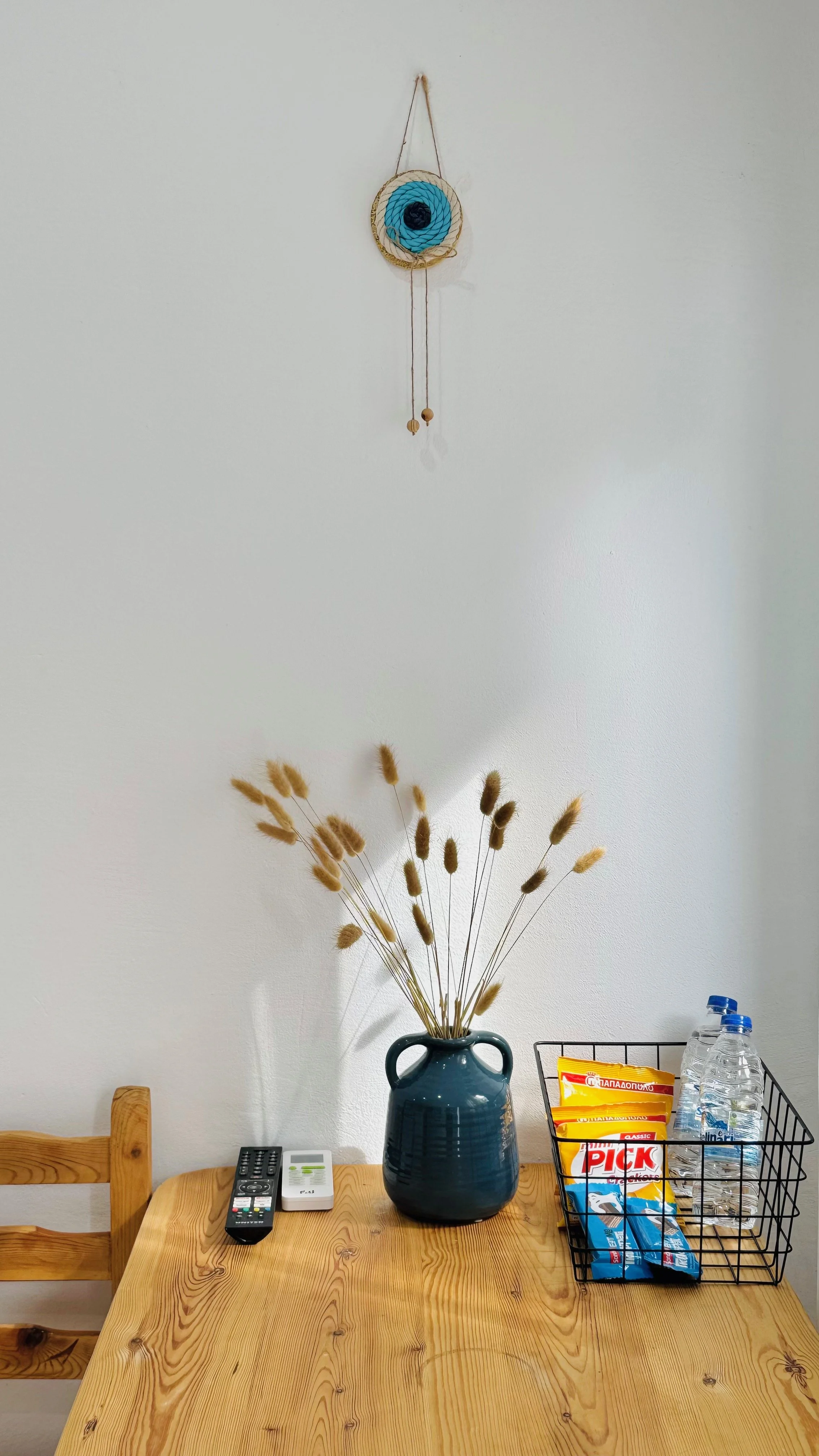 A wooden dining table with a vase of dried grass, a TV remote, a microwave remote, and a wire basket with water bottles and snack packets. A wall-mounted decorative wall hanging with a blue eye design is above the table.
