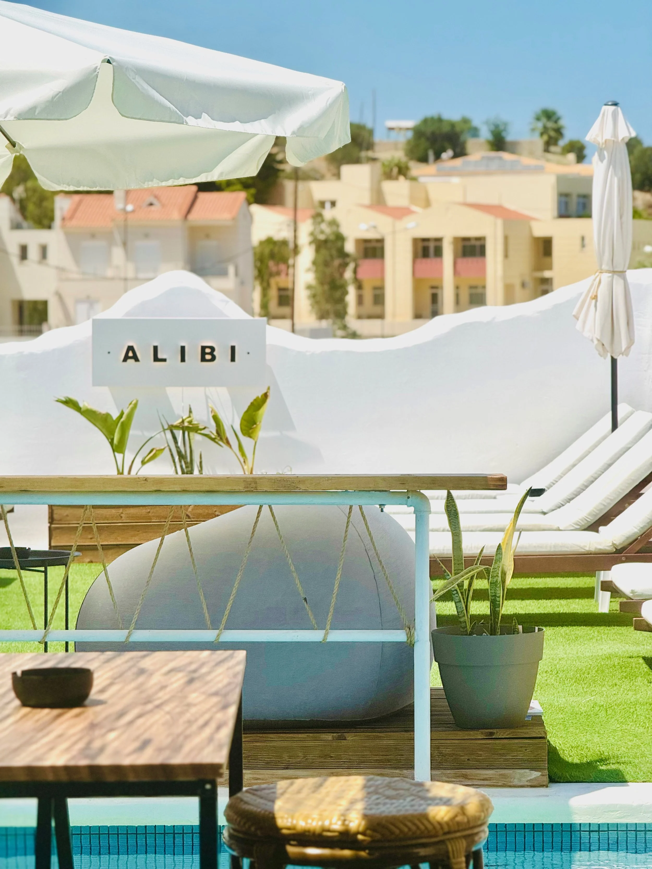 A poolside scene with white lounge chairs, umbrellas, potted plants, and a sign that says 'ALIBI' on a white wall, with buildings and trees in the background on a sunny day.
