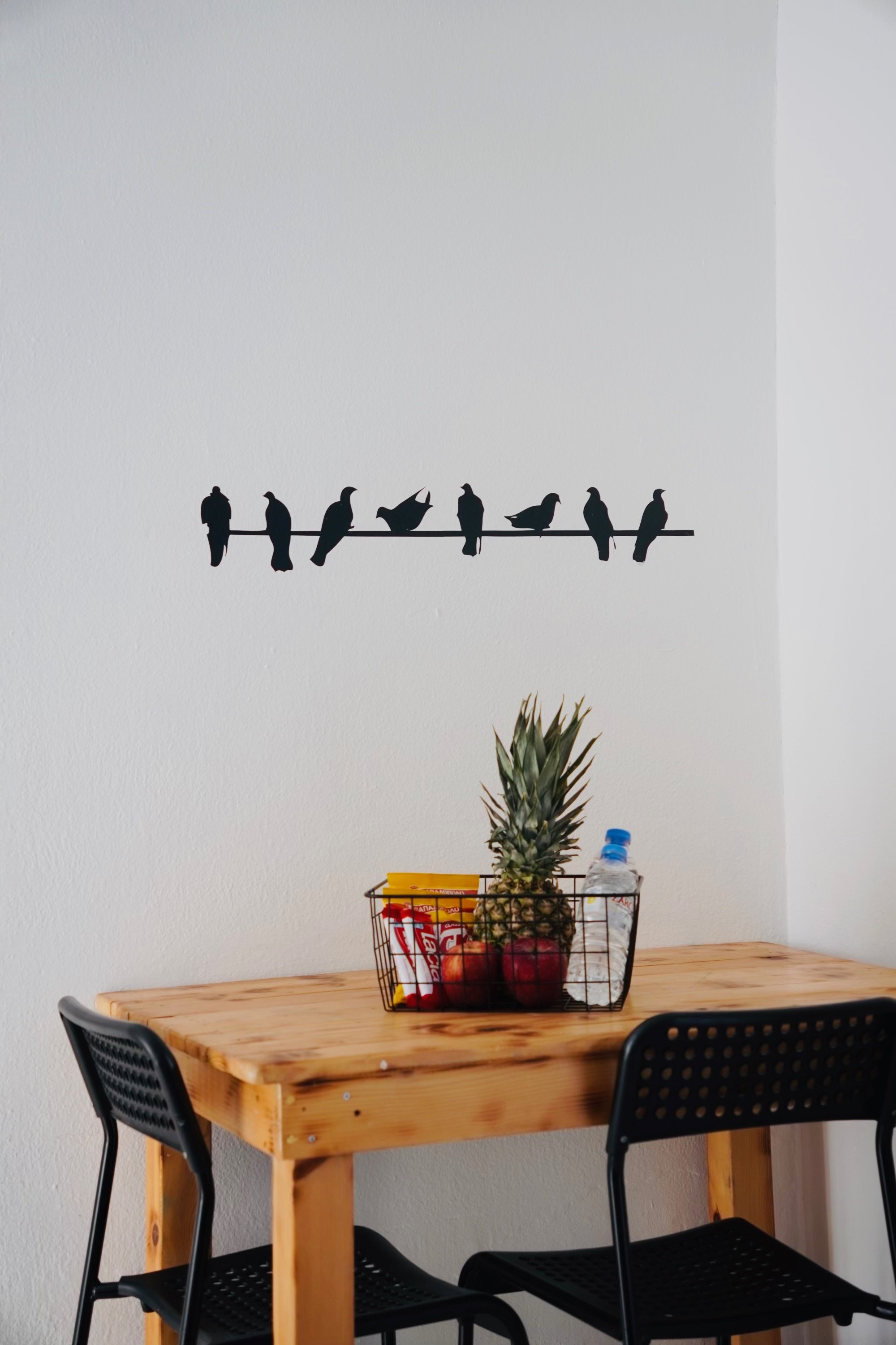 Wooden table with wire basket containing a pineapple, apples, snacks, and a water bottle, two black chairs, and a wall decal of birds on a wire.