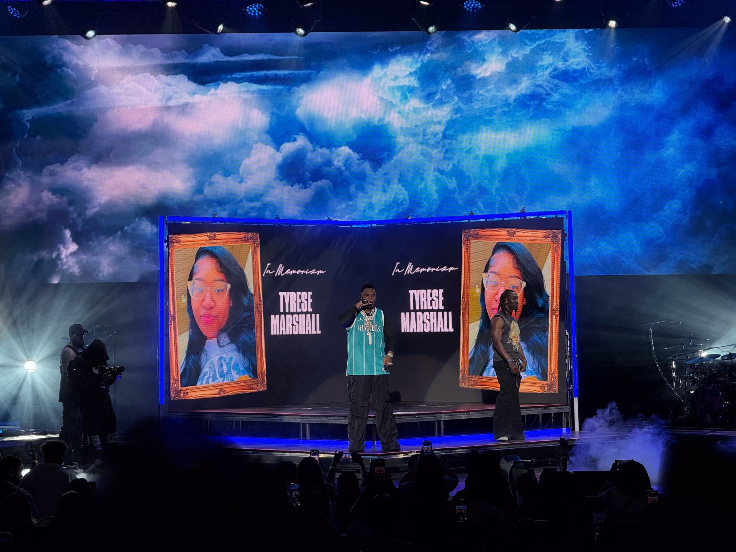 A stage with a large screen displaying an image of a woman with glasses and dark hair, and the text 'In Memoriam Tyrese Marshall'. There is a person speaking into a microphone on stage, with two other individuals standing nearby, and a crowd in front of the stage. The backdrop shows a digital depiction of a cloudy sky.