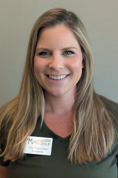 A woman with long blonde hair smiling, wearing a dark green top and a name tag that says "Morningside" and "Cally Ziegenmeyer, Reception" standing against a light-colored wall.