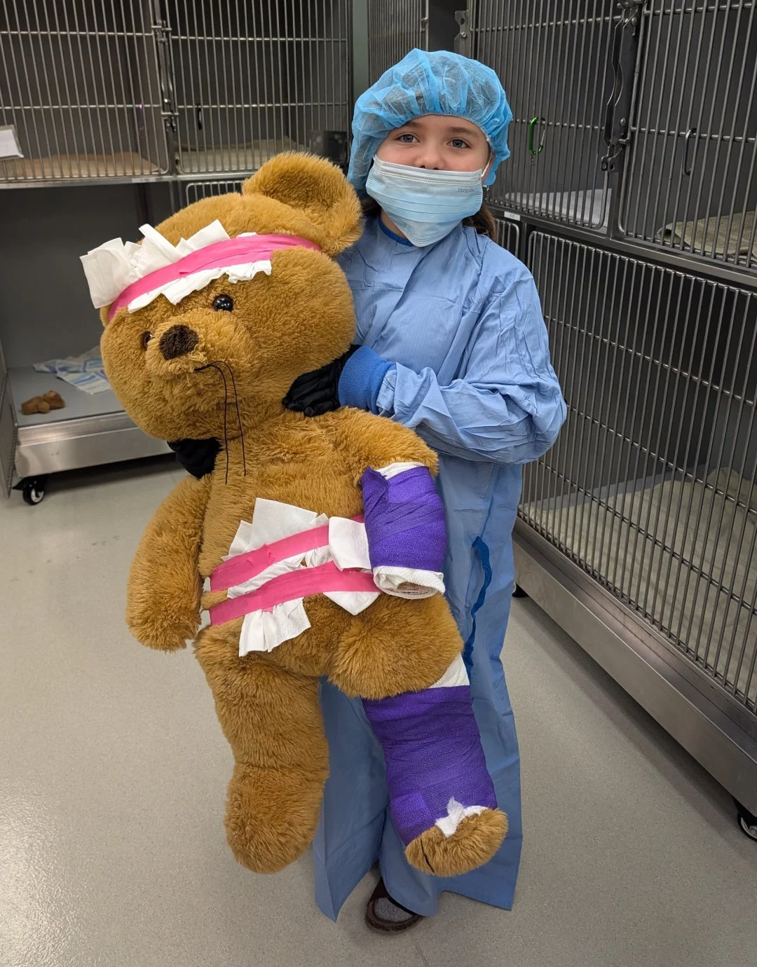 A young girl in medical scrubs, wearing a mask and hair cover, holding a large plush teddy bear with a purple cast on its leg and bandages.