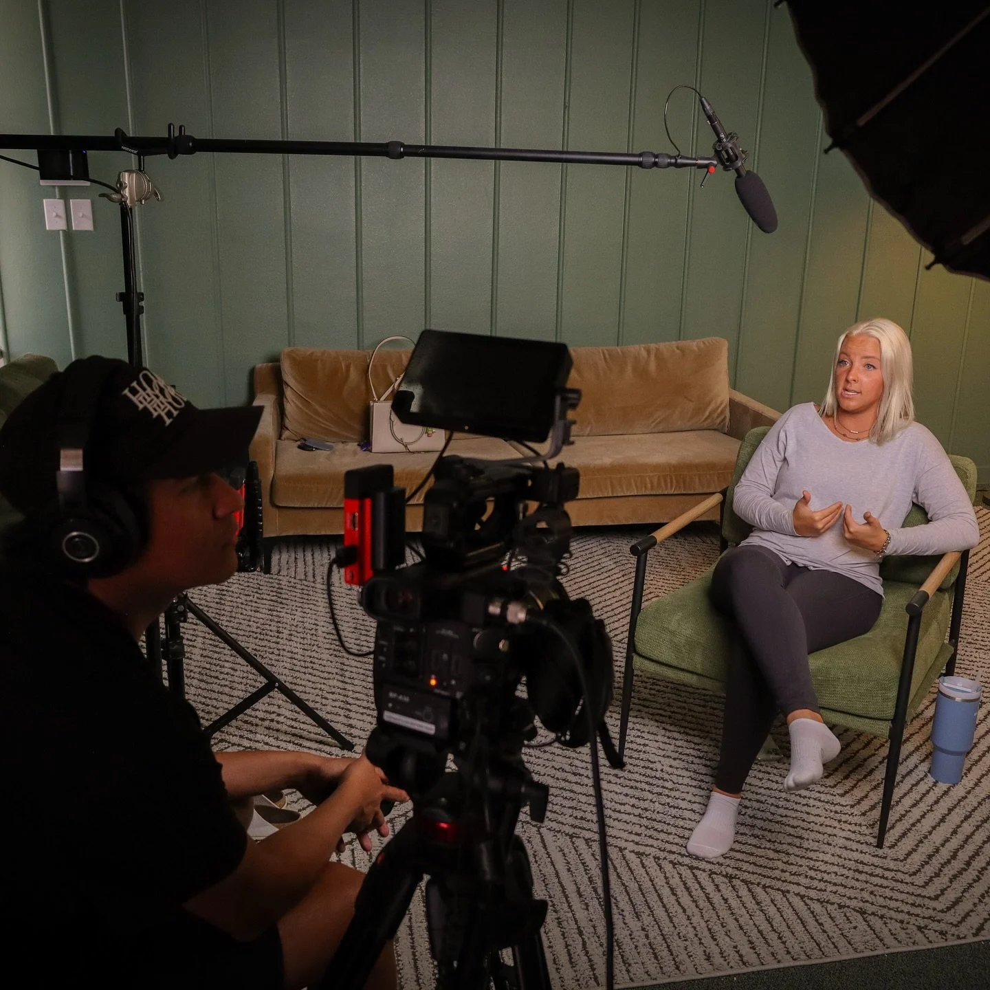 A woman with blonde hair sitting in a green chair during an interview or recording session, speaking and gesturing with her hands. A camera and a sound recorder are capturing the scene, with a person in headphones operating the camera. A beige couch, a bag, and a blue mug are visible in the background. The room has green walls and a patterned carpet.