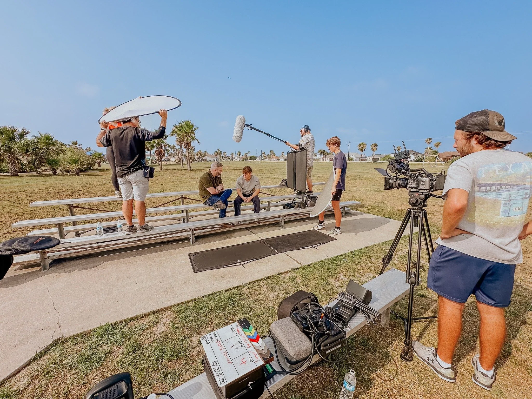 Film crew filming a conversation on bleachers in a park with palm trees under a blue sky. Crew members are managing equipment, including a camera on a tripod and an audio boom microphone.
