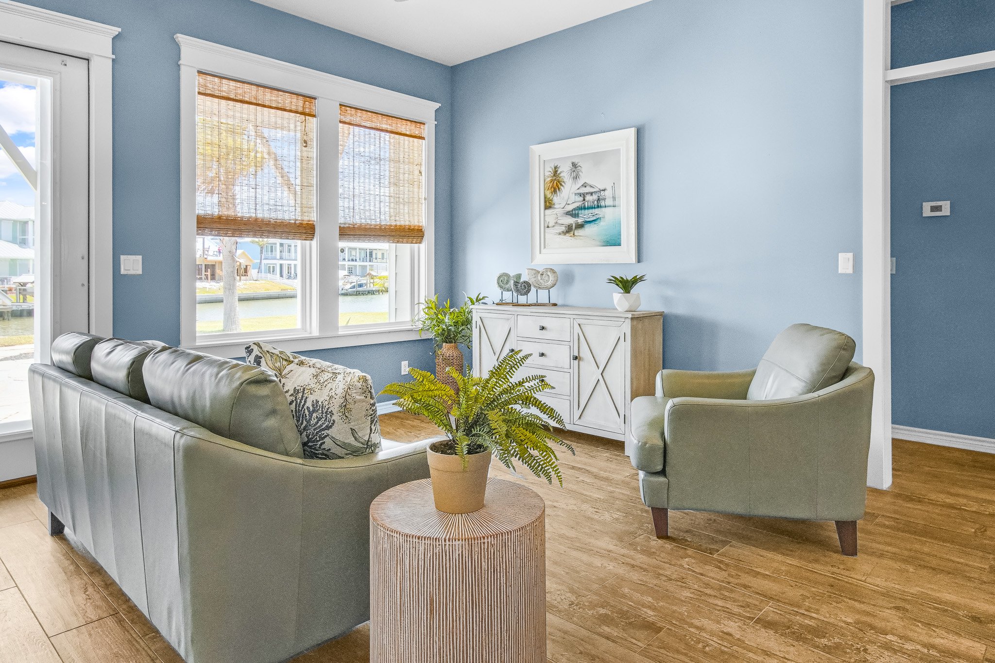 Living room with light blue walls, large windows with bamboo blinds, a gray sofa with patterned pillows, a green armchair, a circular wooden side table with a potted fern, and a white sideboard with decor and a framed artwork of a beach scene.