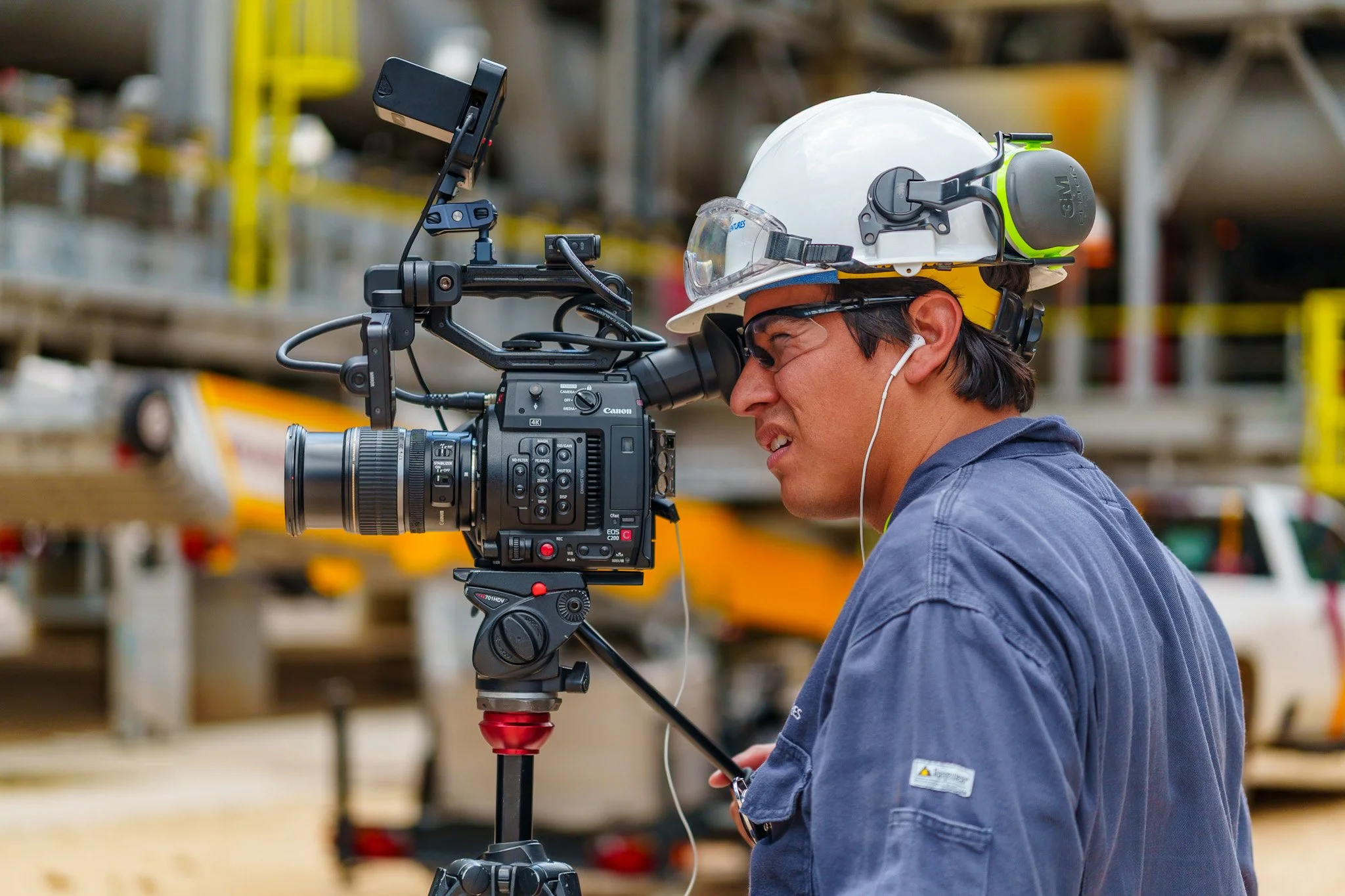 A man in a blue jacket, safety glasses, white hard hat, and hearing protection filming with a professional video camera on a tripod at an industrial site.