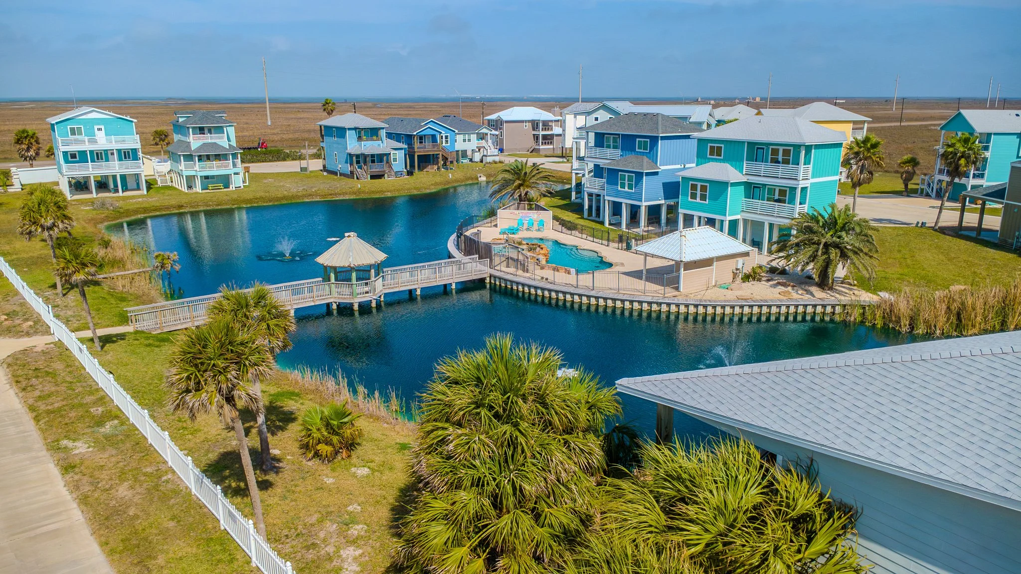Colorful beach houses around a pond with a fountain and palm trees on a sunny day.