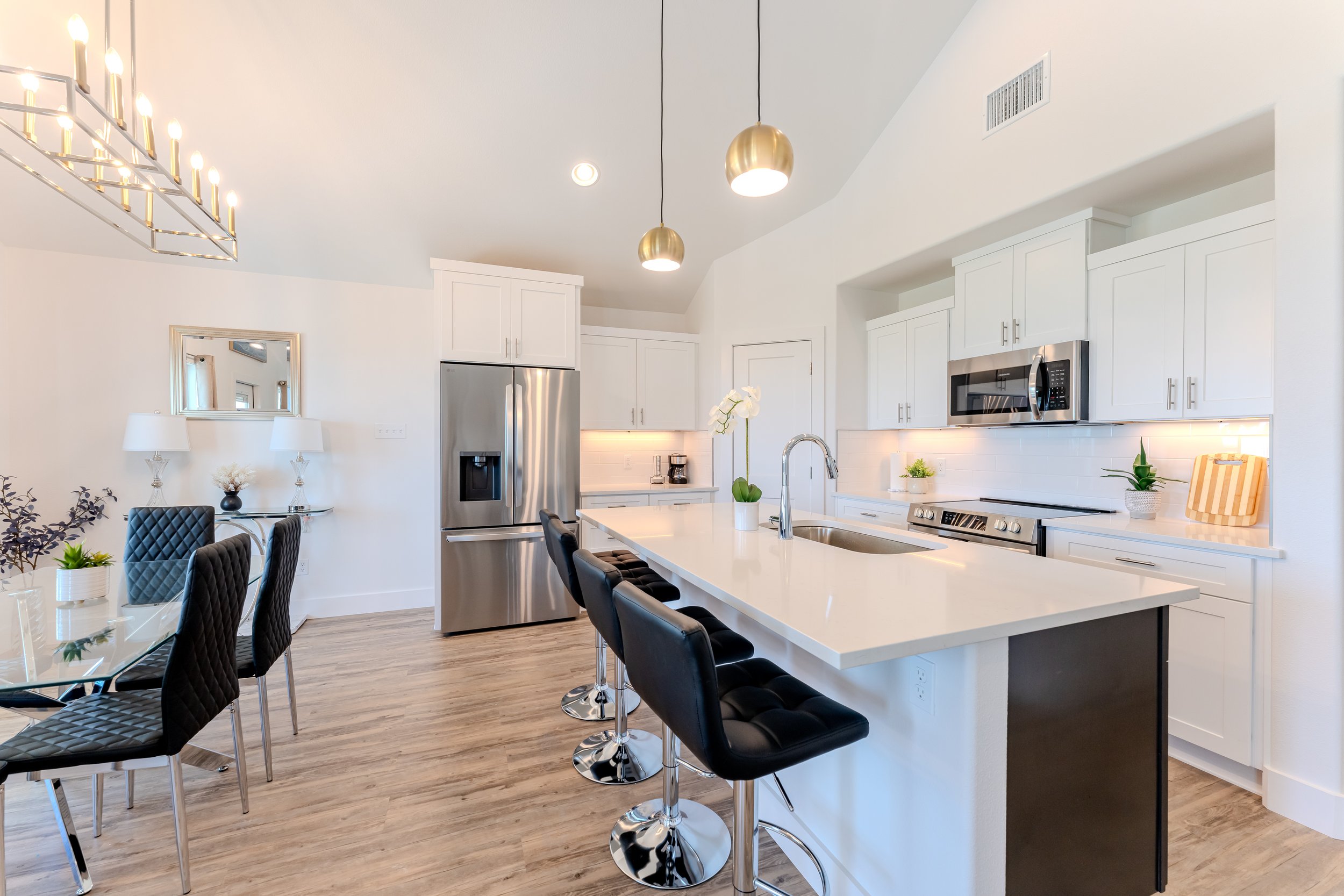 Modern kitchen with white cabinets, stainless steel appliances, and a large island with black barstools; adjacent dining area with glass table and black chairs.