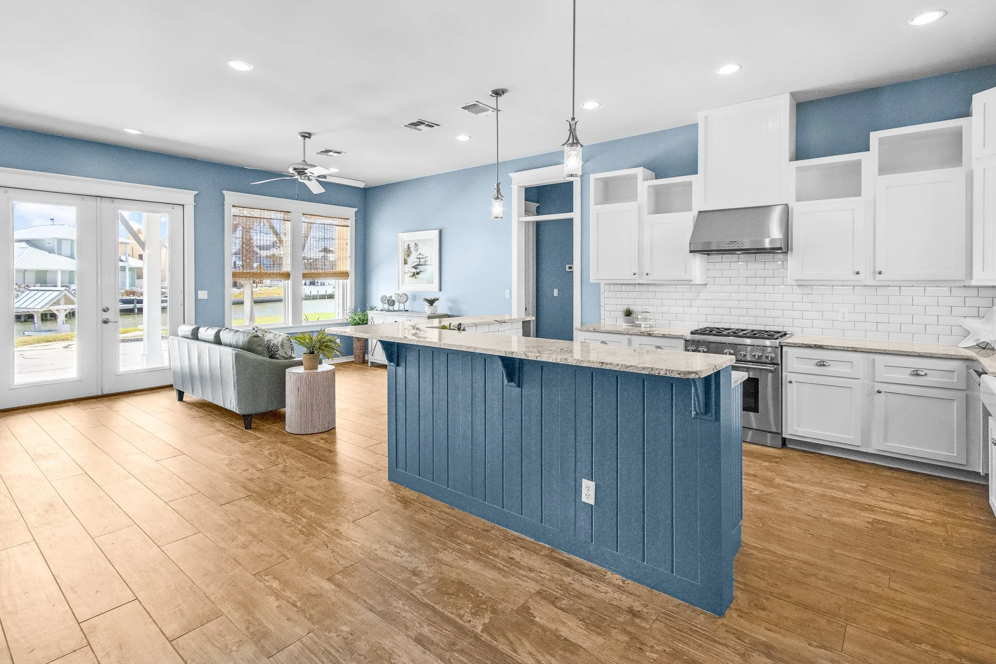Open-concept kitchen and living room with blue walls, white cabinets, stainless steel appliances, light granite countertops, hardwood floors, and large windows with blinds. There's a light blue sofa and small side table near the windows.