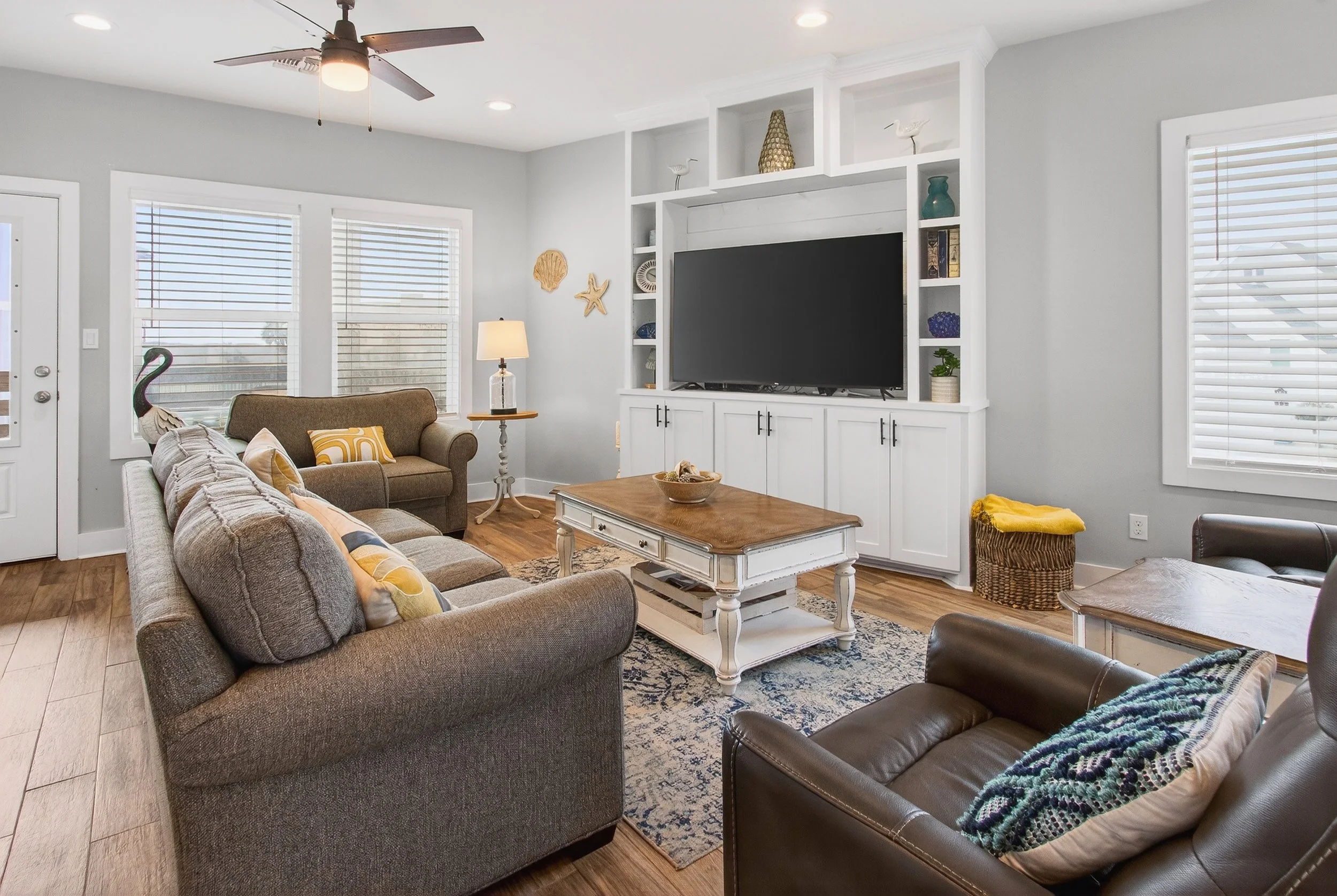 Living room with a gray sofa, brown armchair, wooden coffee table, white entertainment center with a large TV, white shelves with decorations, large windows with blinds, ceiling fan, and a lamp on a side table.